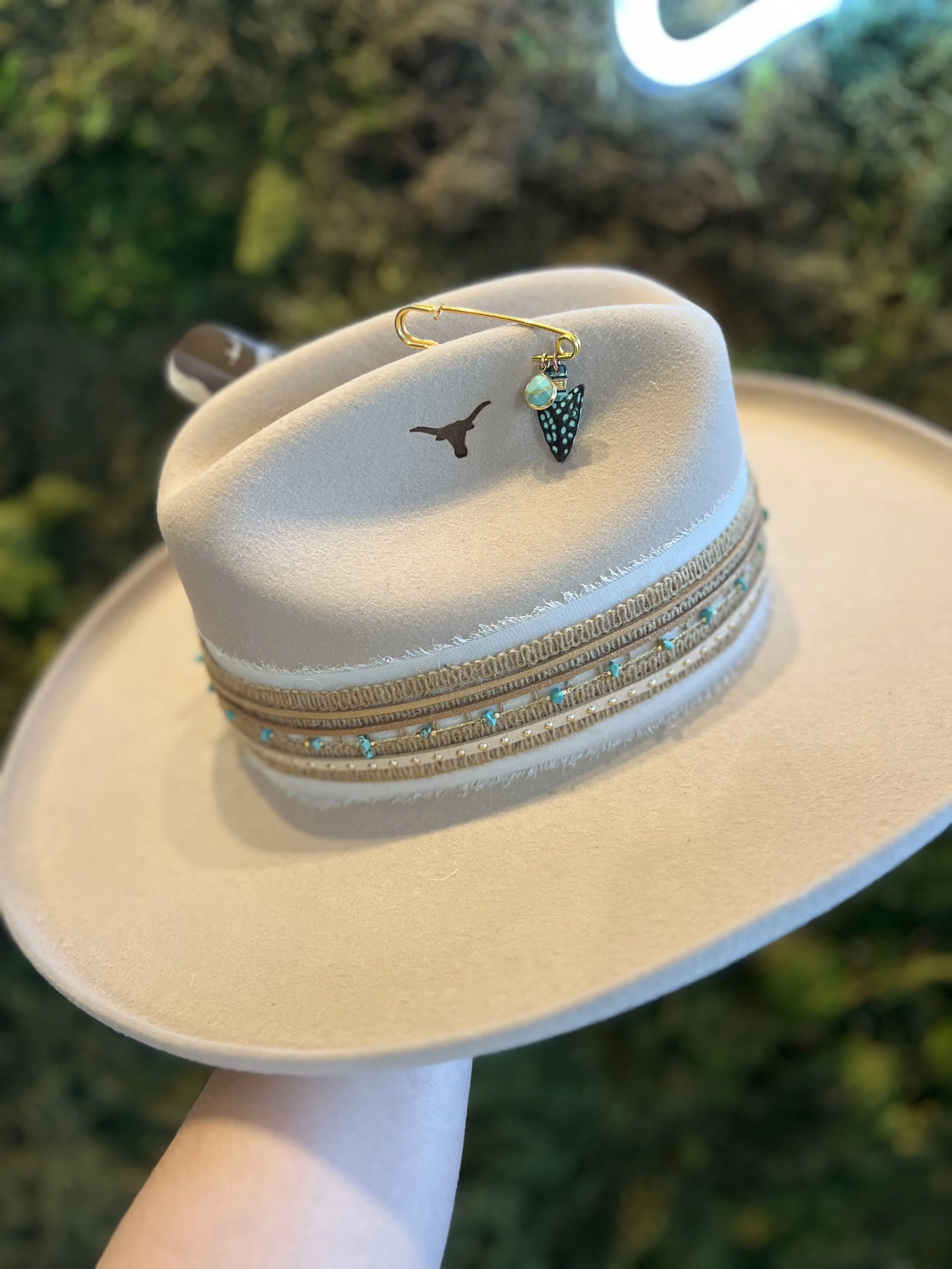 A gold safety pin with a black and turquoise butterfly charm and a small pearl charm attached, placed on a light beige hat decorated with turquoise beads and gold accents, outdoors with a green, blurry background.
