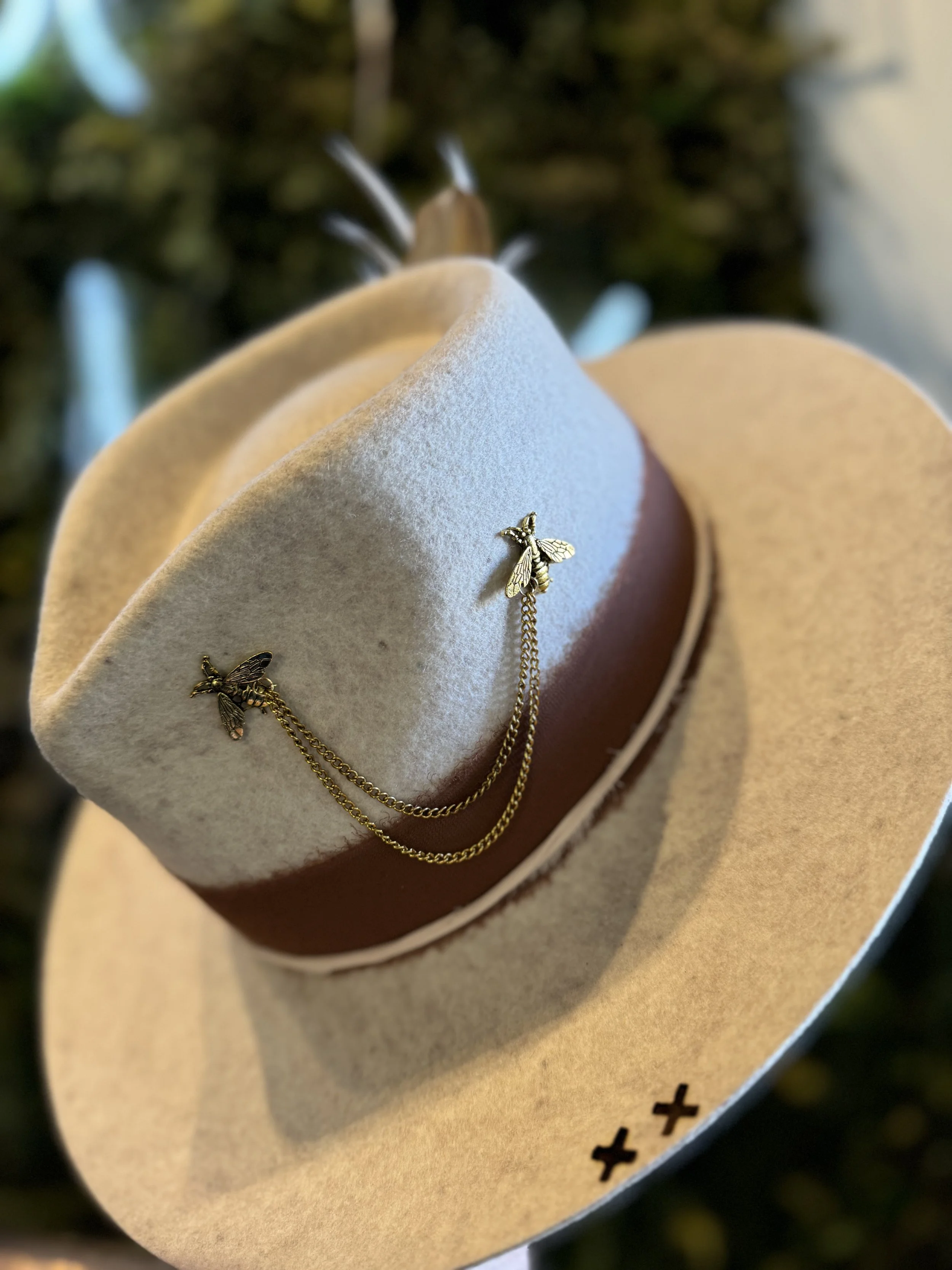A beige wide-brimmed hat with a brown band, decorated with gold bee pin and chains, placed on a blurred background with lights.