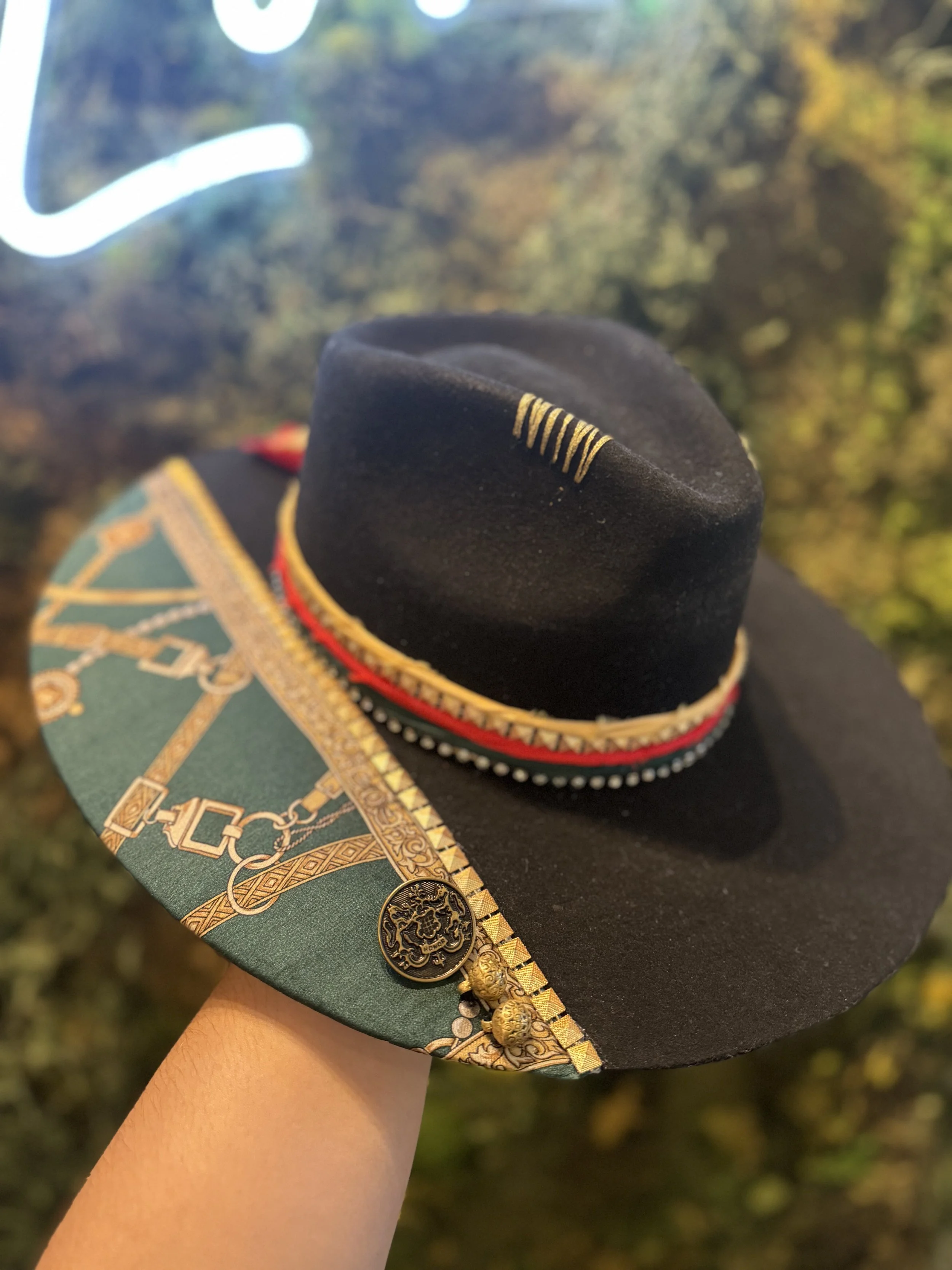 A person's arm holding a decorated black felt hat with embroidered designs, gold embroidery, and jewels, against a blurred background of trees with fall foliage.