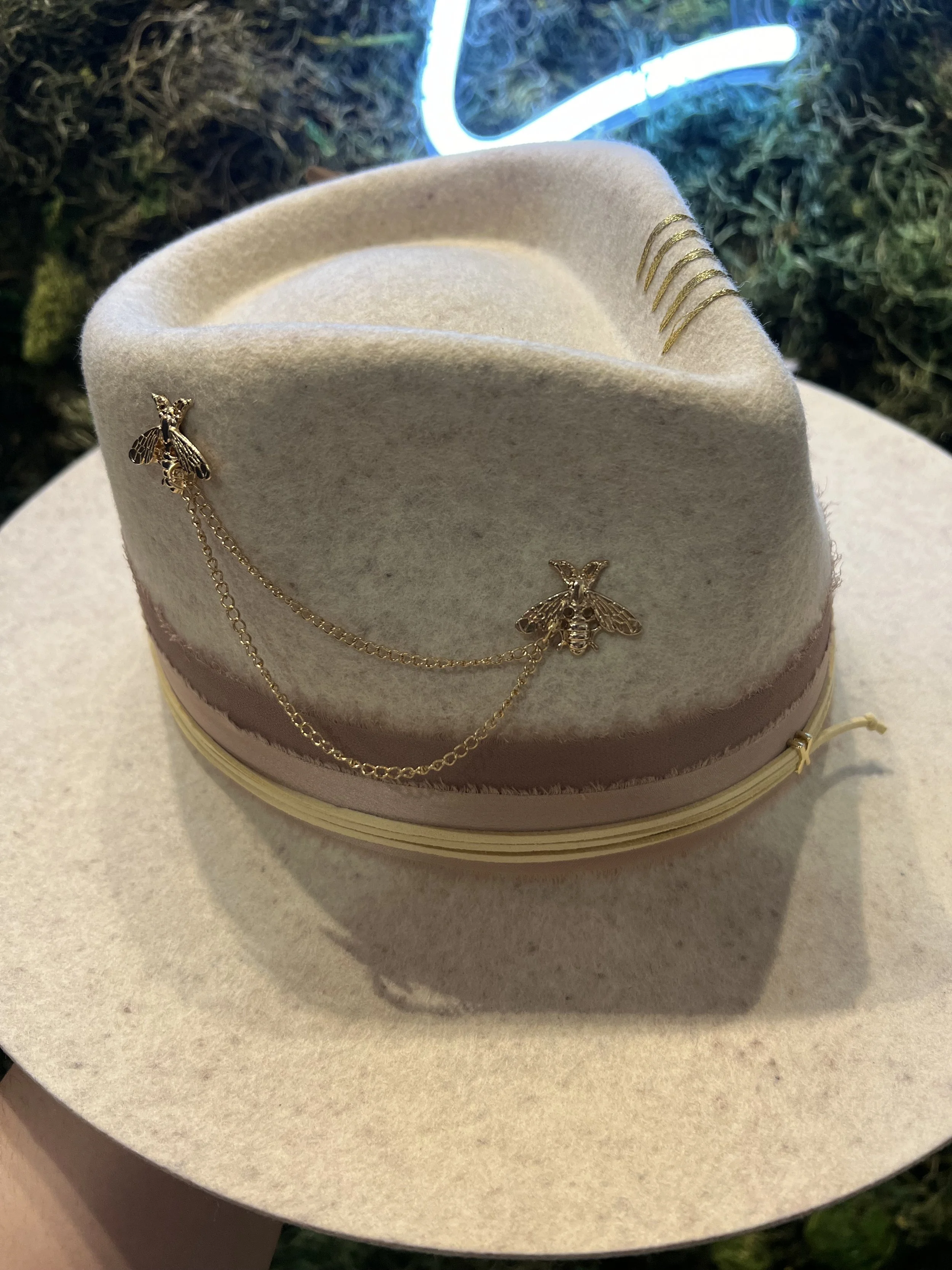 A cream-colored felt hat decorated with gold bee and chain embellishments, set against a nondescript background.