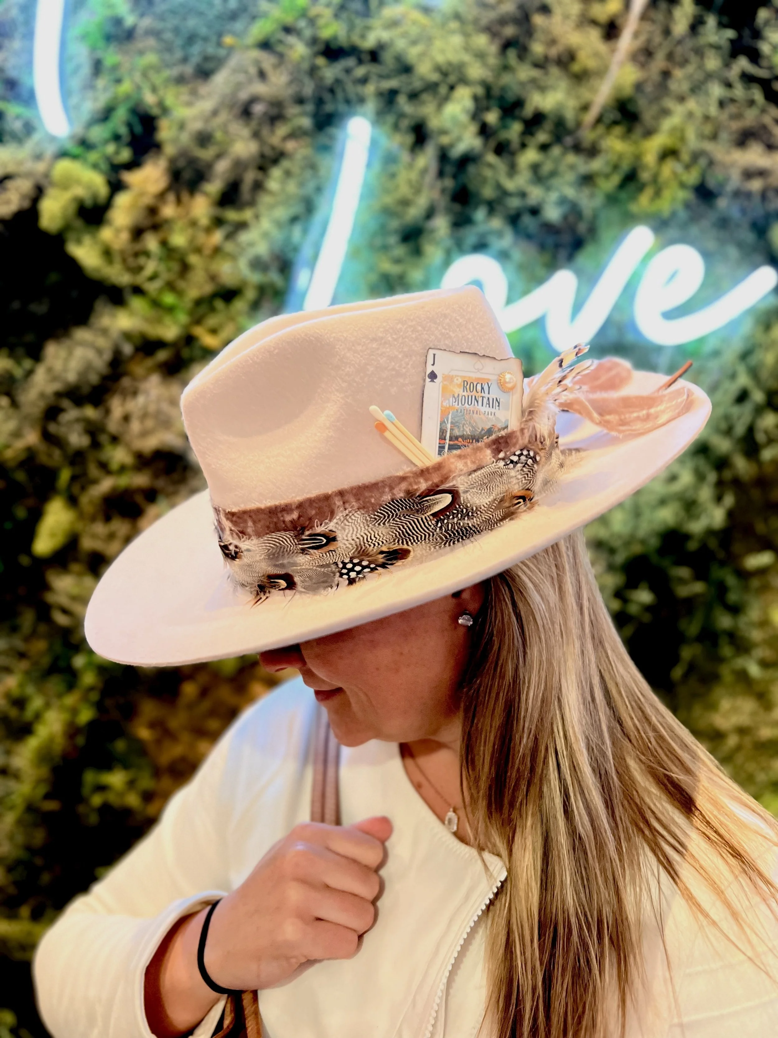 A woman wearing a white felt hat decorated with feathers and a playing card, standing outdoors with greenery and a neon 'love' sign in the background.