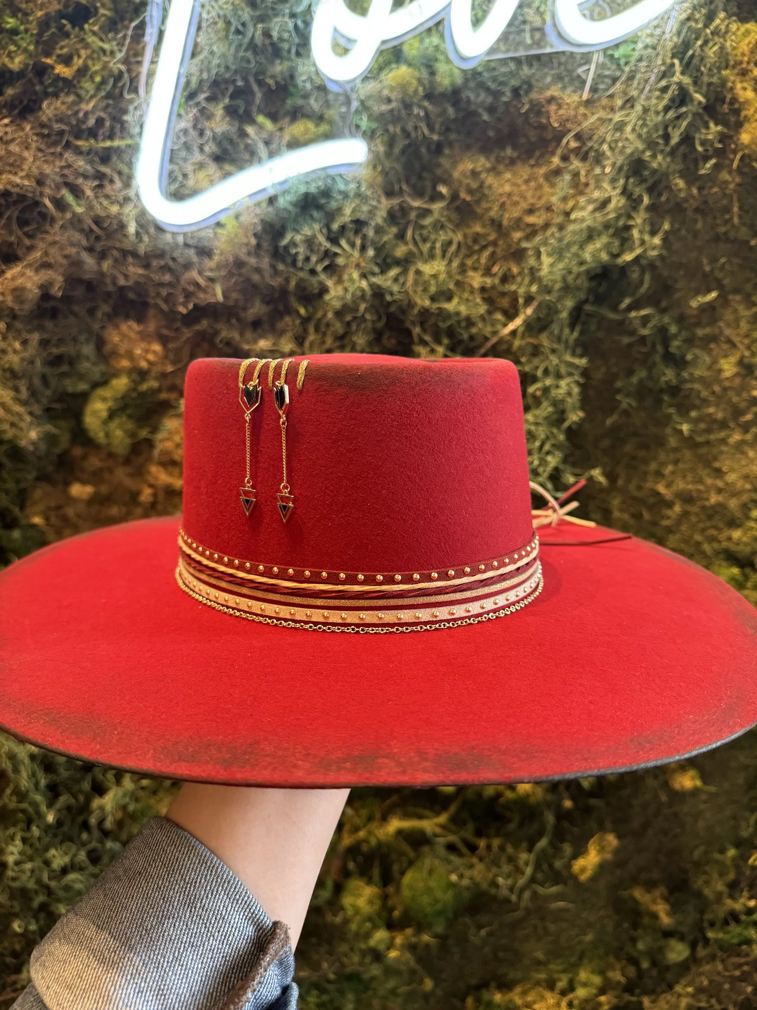 A red felt hat decorated with several layered bands of gold and silver chains, with a pair of gold earrings hanging from the top band, held up against a background of moss and a neon light.