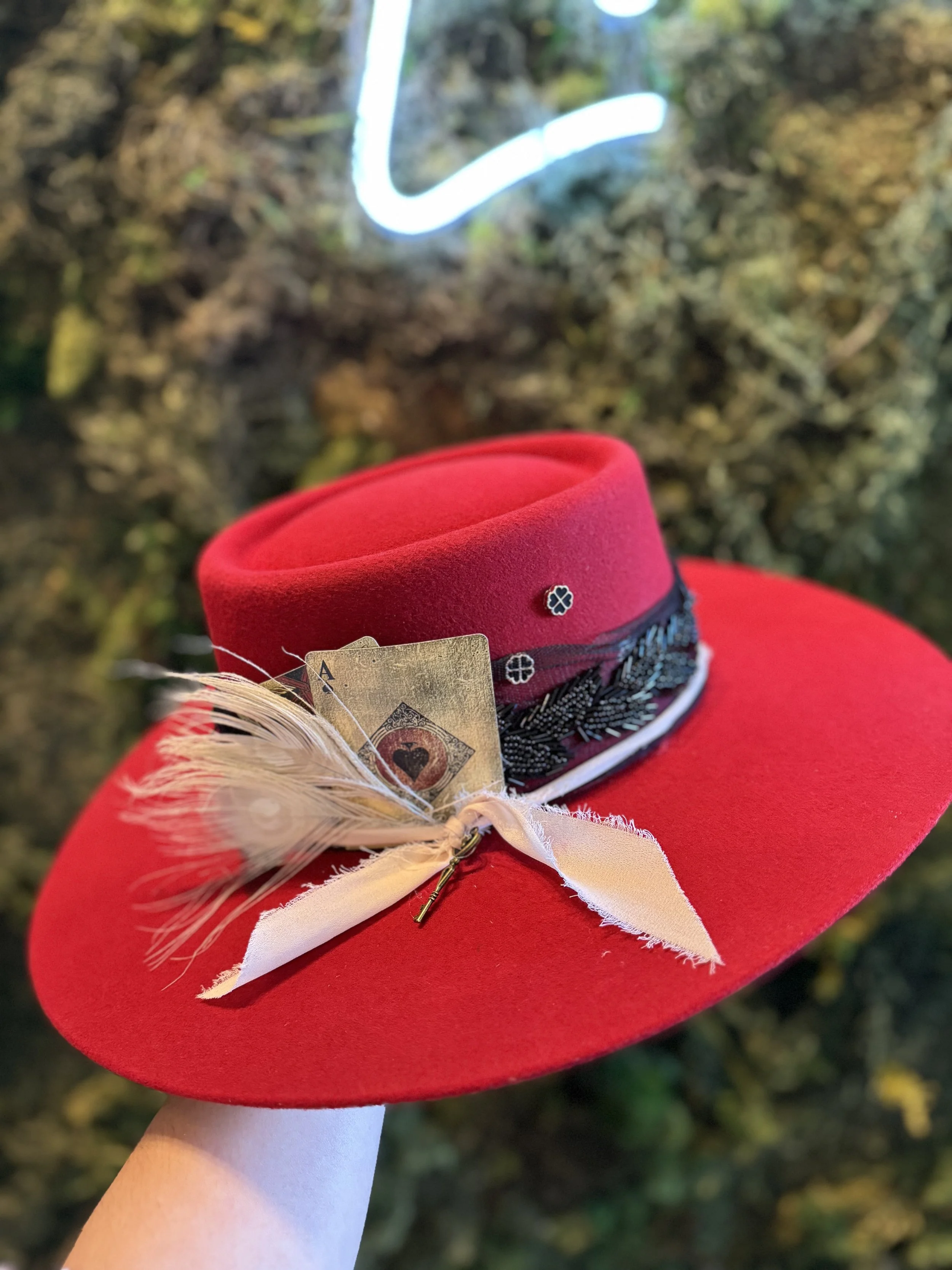 A red felt hat with decorative elements, including a cannabis leaf pattern band, a feather, an ace of spades card, and a small key, held by a person in front of a blurred outdoor background.