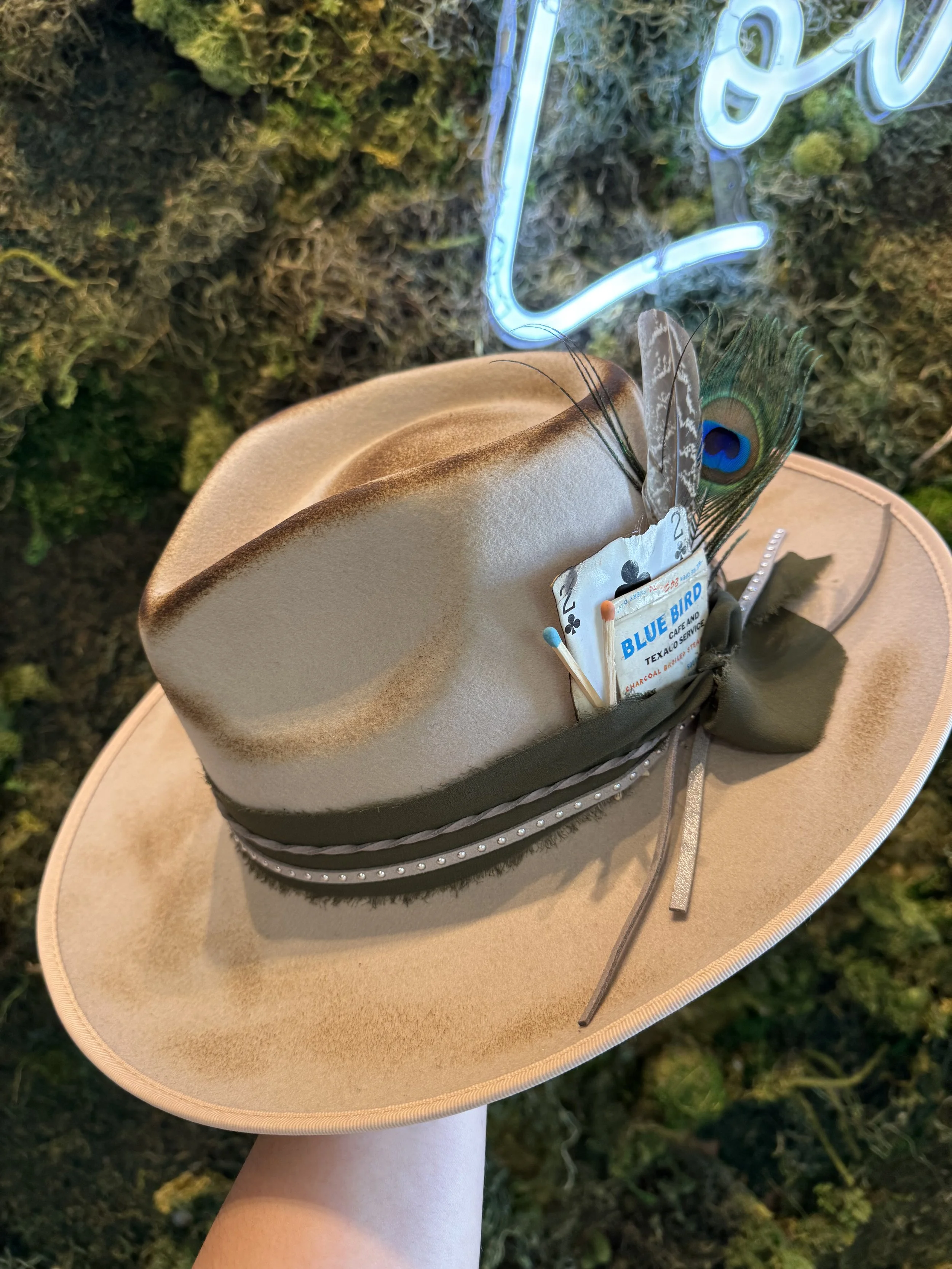 A cowboy hat with a decorative band, decorative pins, a peacock feather, a pack of Blue Bird cigarettes, and a playing card, held against a background of moss and a neon sign.