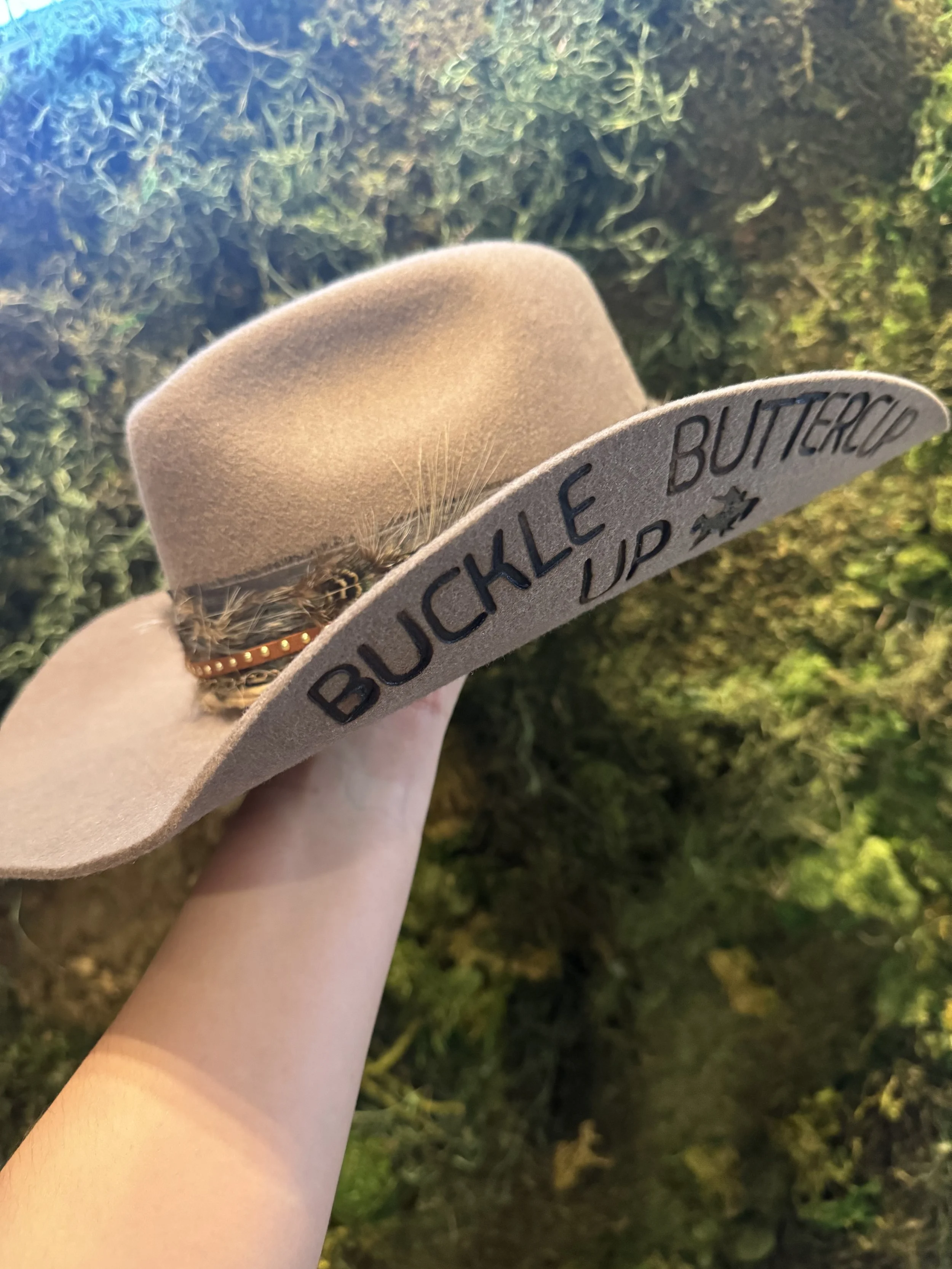 A beige felt hat with a feather, being held outdoors near greenery. The brim of the hat has the words "BUCKLE UP BUTTERCUP" written on it.