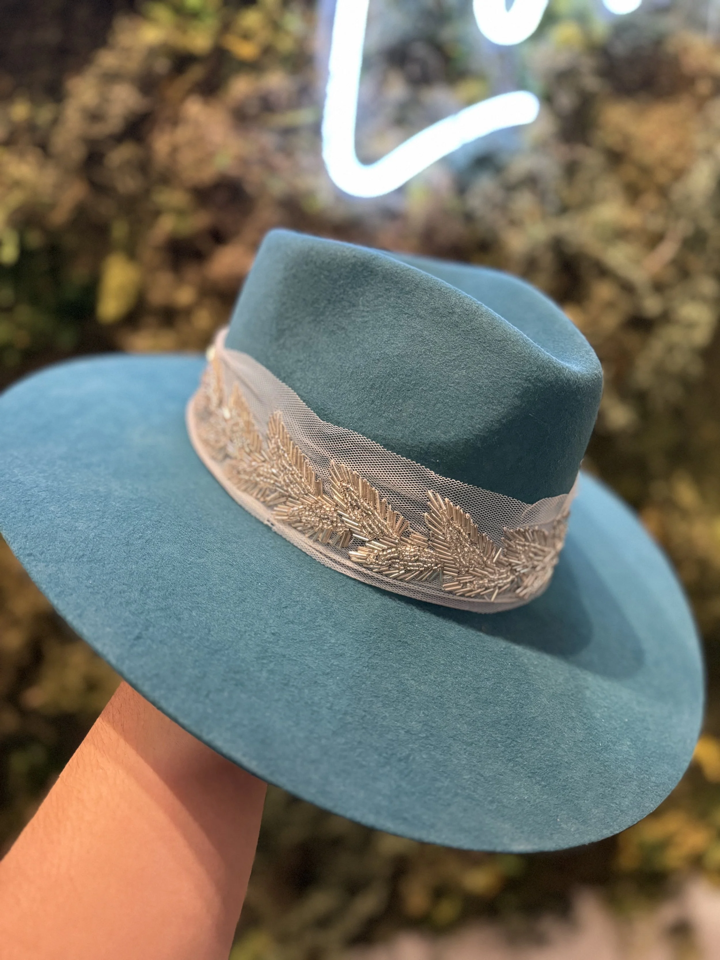 A person holding a blue felt hat with a decorative band of gold embroidery and mesh, against a blurred background of foliage and a neon sign.