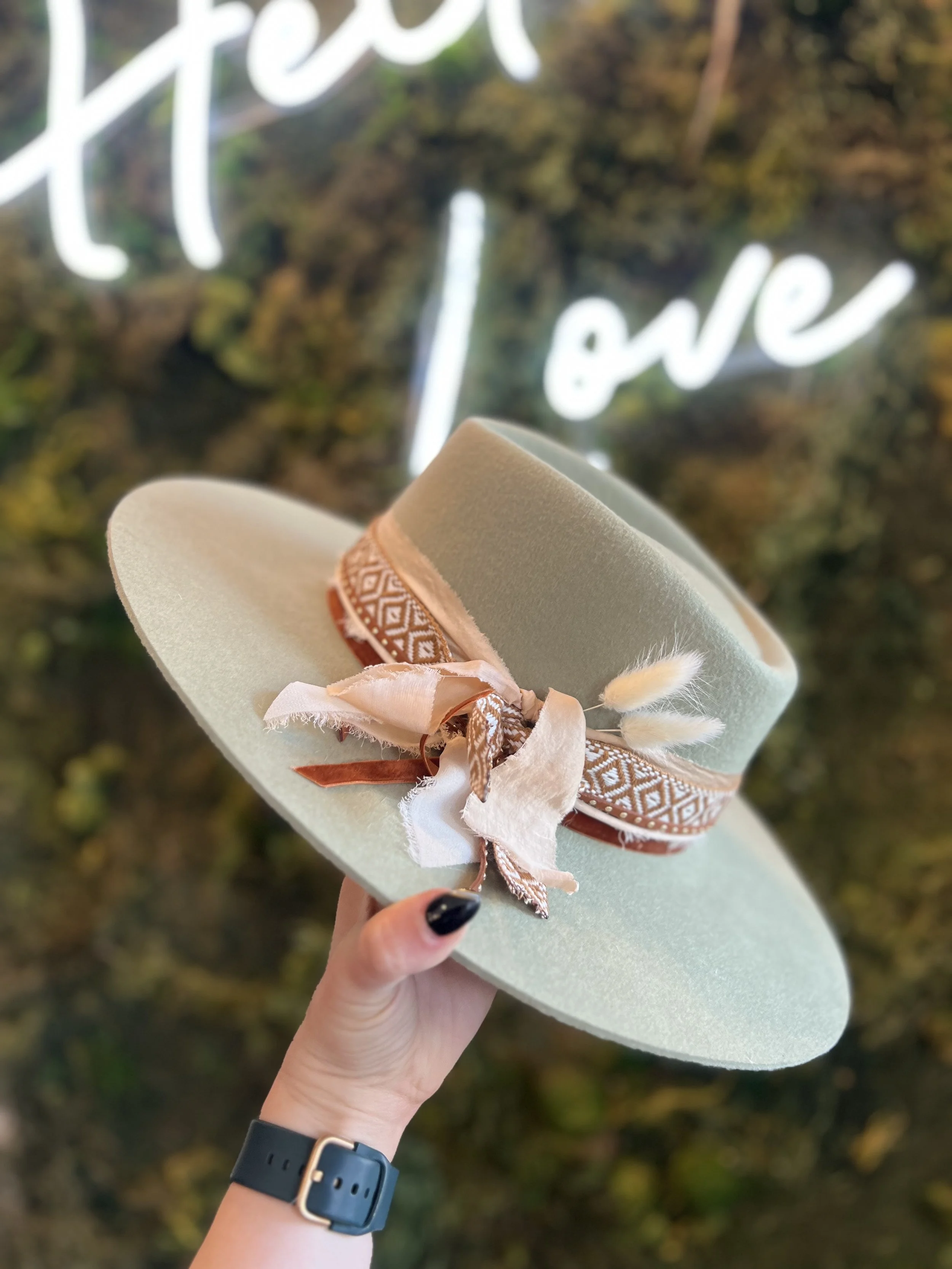 A hand holding a beige wide-brim hat decorated with a patterned ribbon and fabric bow, with a blurred background featuring the words 'Peace' and 'Love' written in white neon lights.