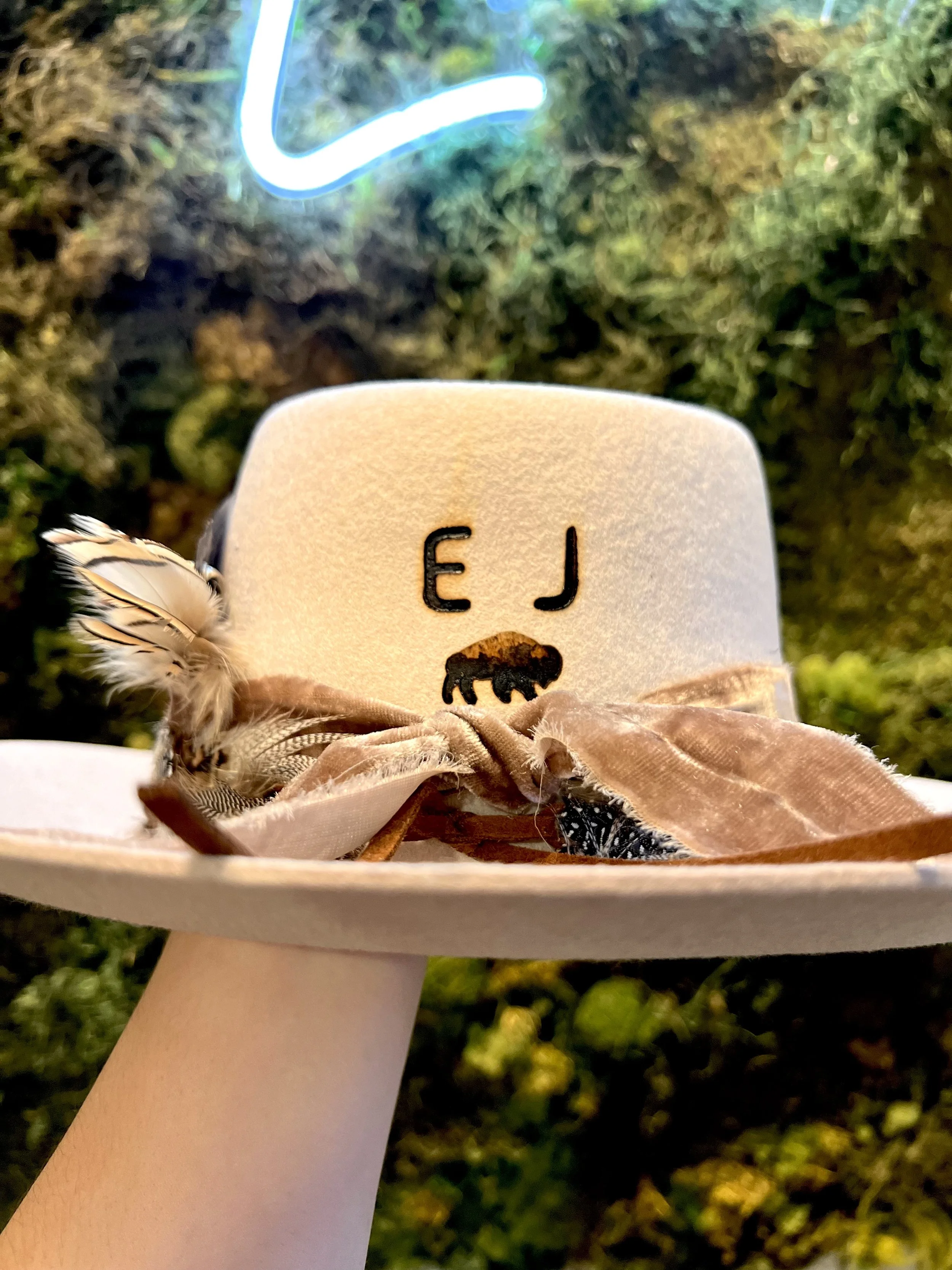A beige hat with a black bear silhouette and the letters 'E J' on the front, decorated with feathers and ribbons, being held up against a background of green moss and illuminated by a neon light.