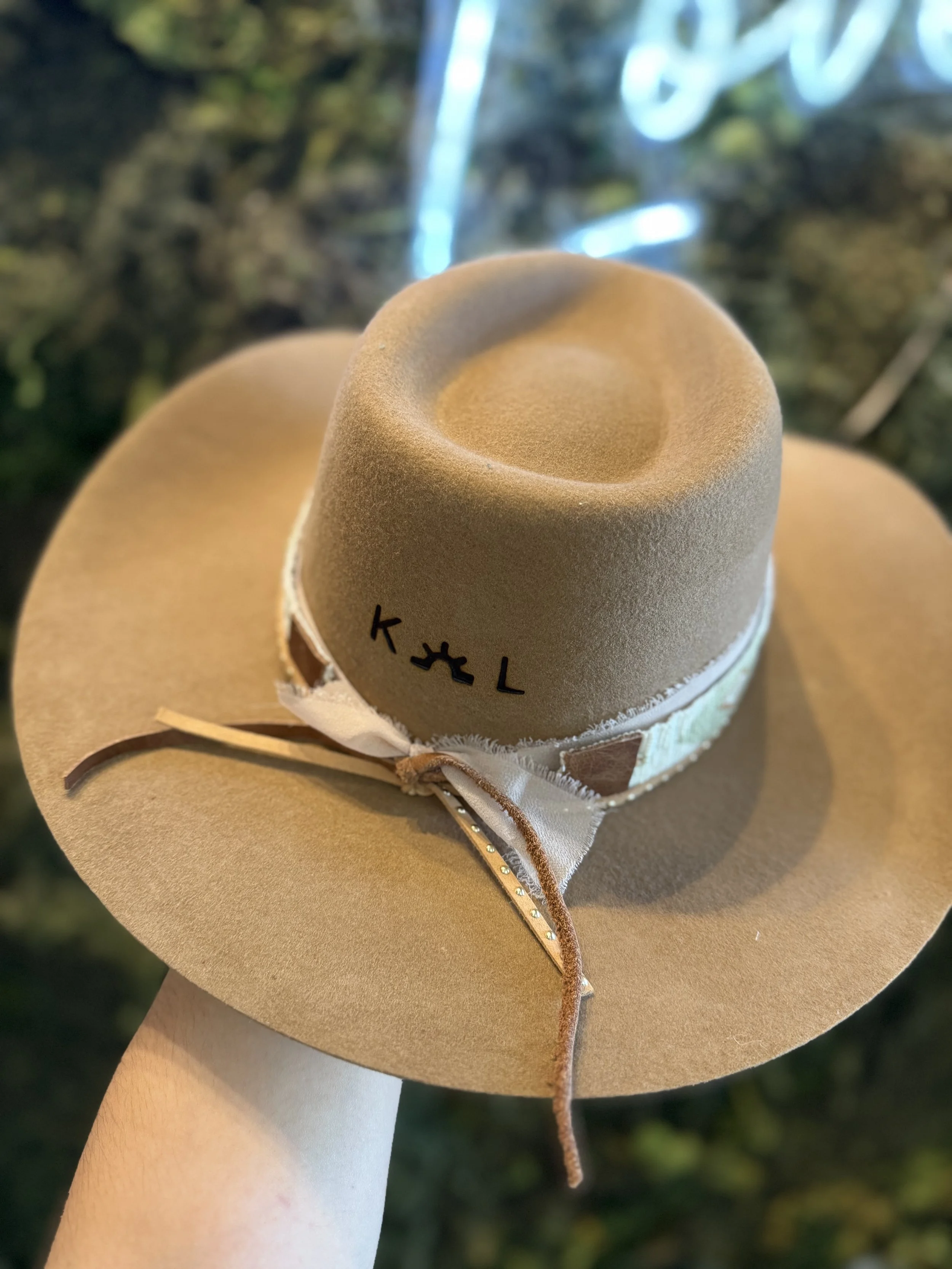 A tan cowboy hat with the initials 'K L' embroidered on it, decorated with a ribbon and small embellishments, held up against a blurred background of trees and a waterfall.