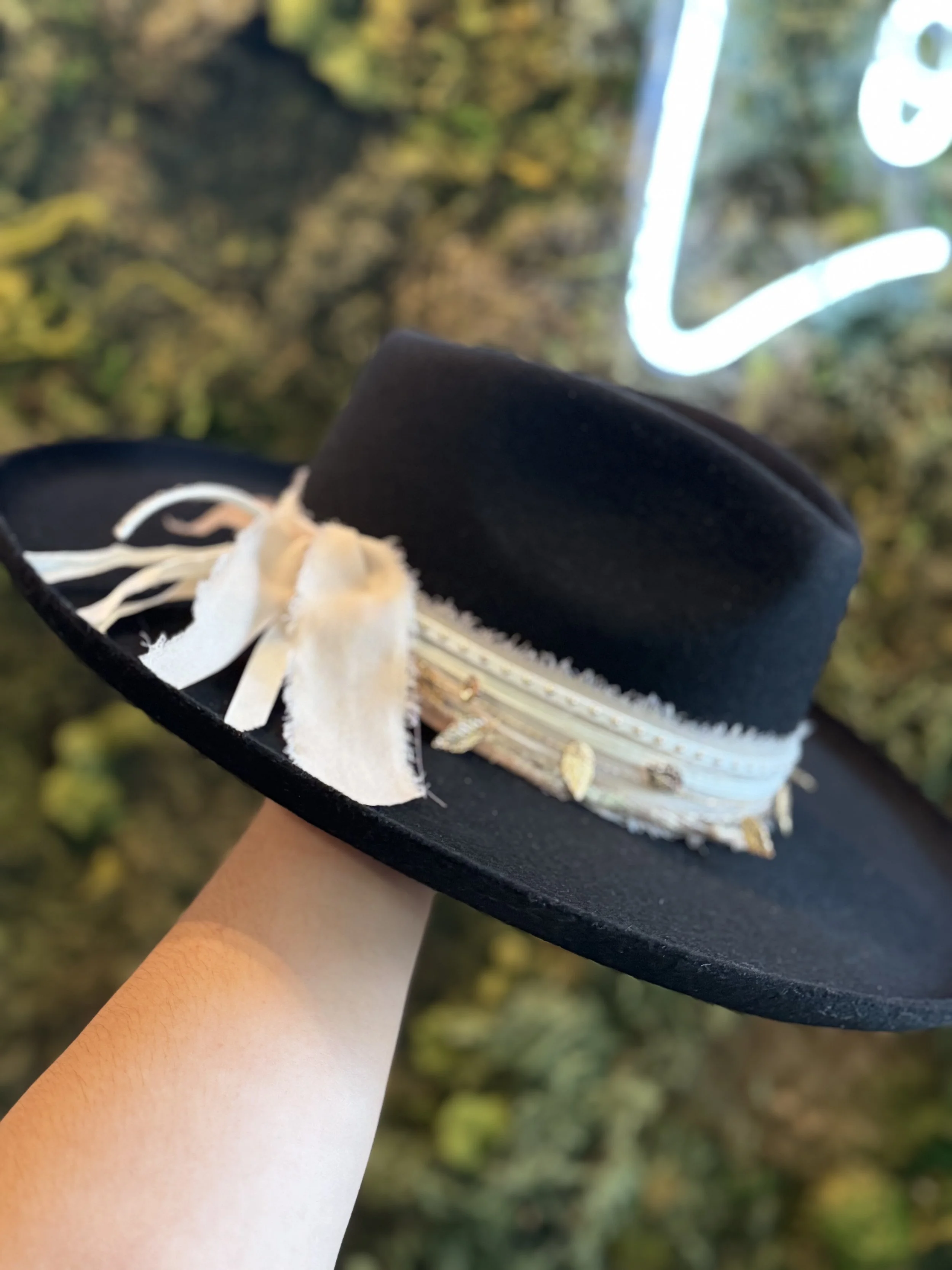 Black wide-brimmed hat with decorative band and white ribbon, held by a person with light skin, against a blurry outdoor background with green foliage and a neon sign.