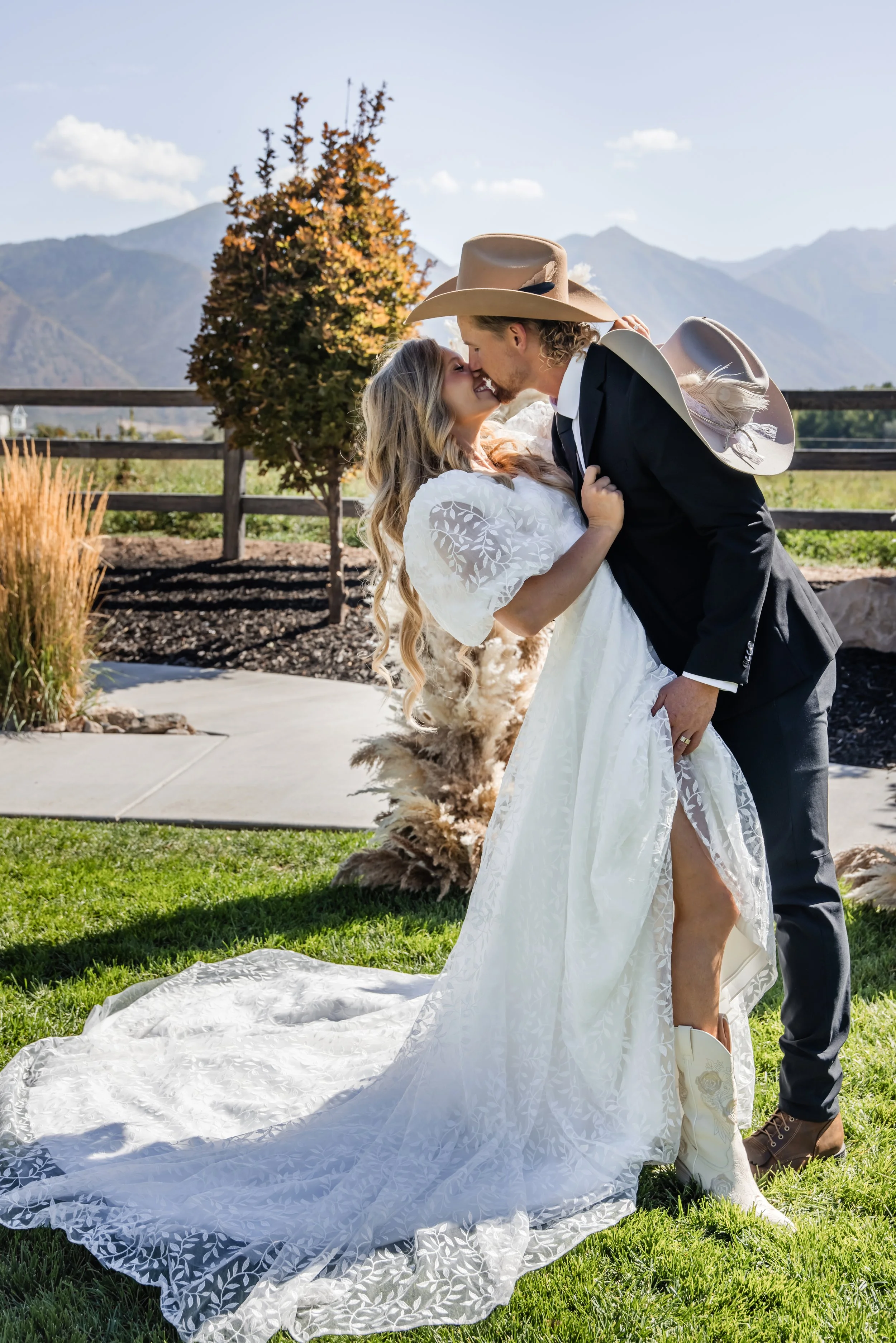 A couple dressed in wedding attire sharing a kiss outdoors, with mountains and a fence in the background.
