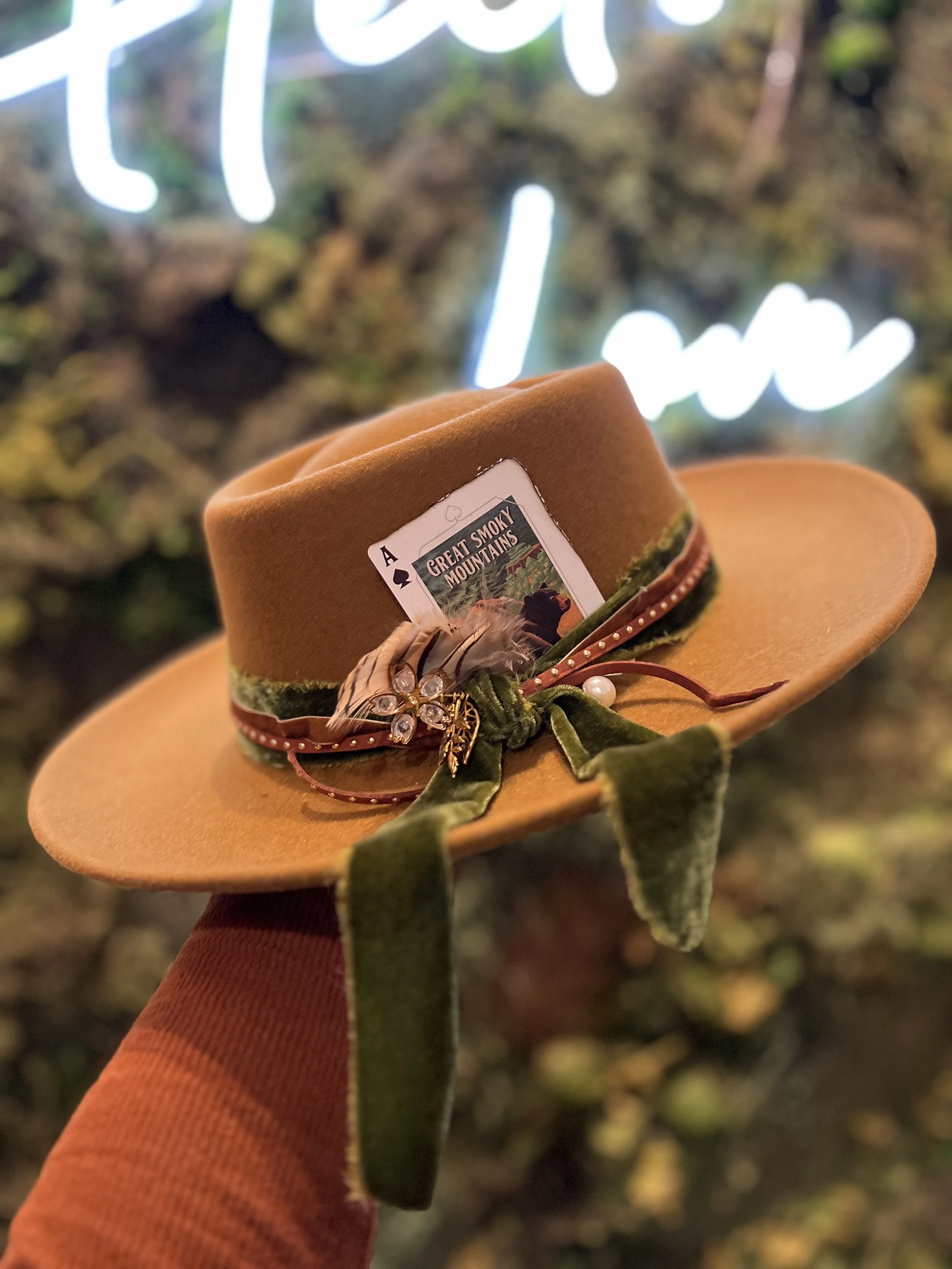 A brown felt hat decorated with a vintage playing card, a feather, a pearl, a brooch, and a green ribbon. The hat is held against a blurred background with greenery and a lit-up neon sign.