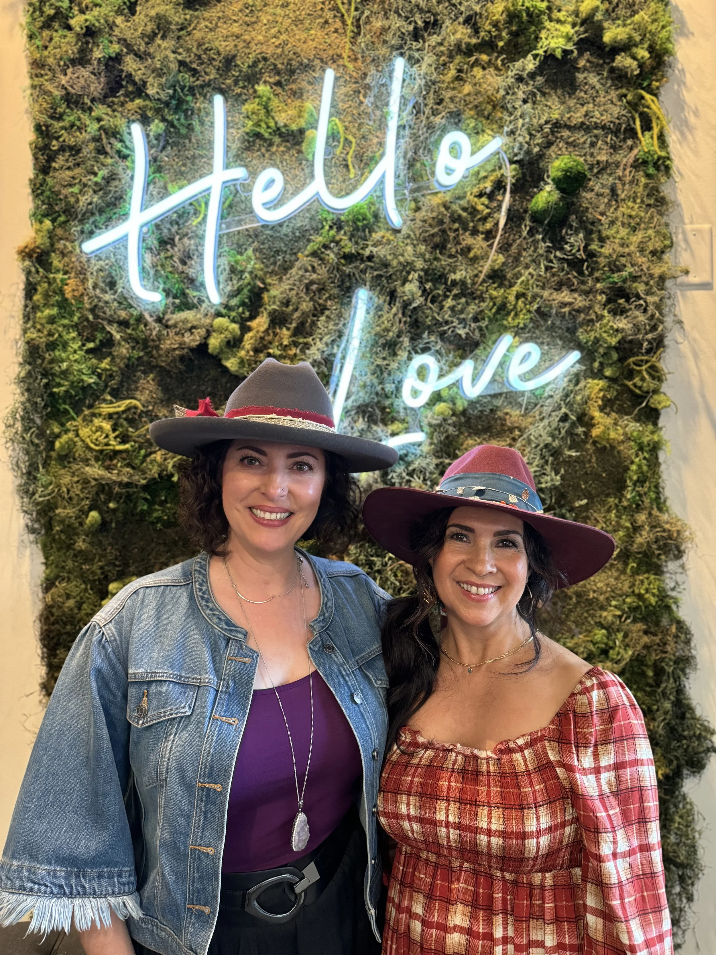 Two women smiling in front of a wall with green moss and a neon sign that reads 'Hello Love'. One is wearing a gray wide-brimmed hat and a denim jacket, the other is wearing a red and white plaid dress and a maroon wide-brimmed hat.