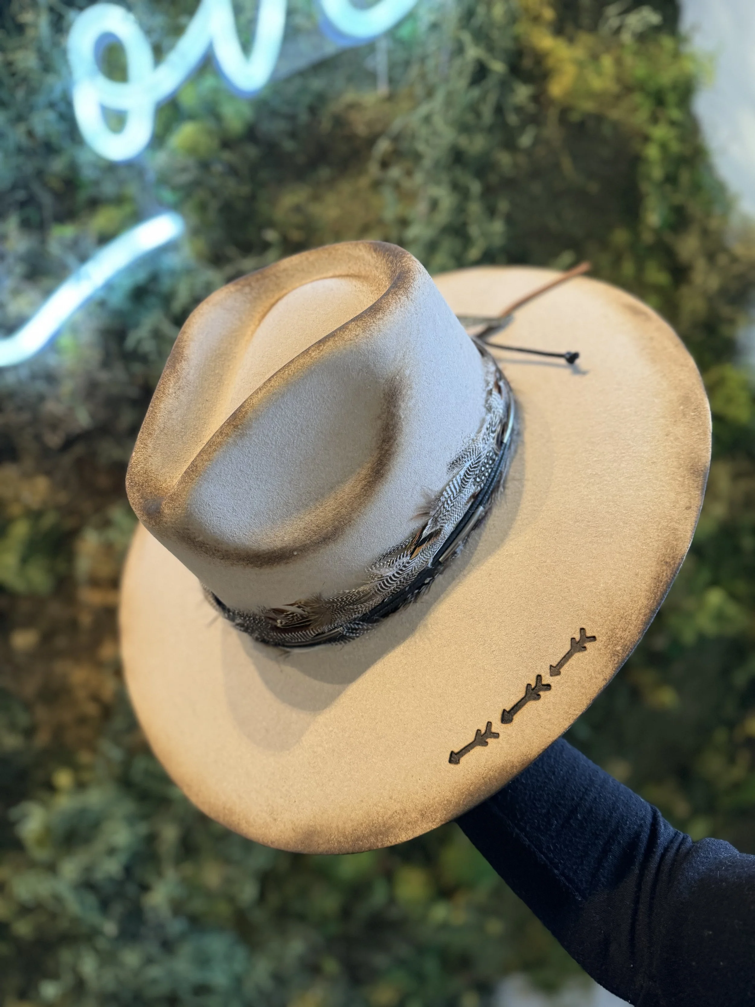 A tan cowboy hat with decorative black and white band, held by a person wearing a black sleeve, with a background featuring green foliage and a blue neon sign.