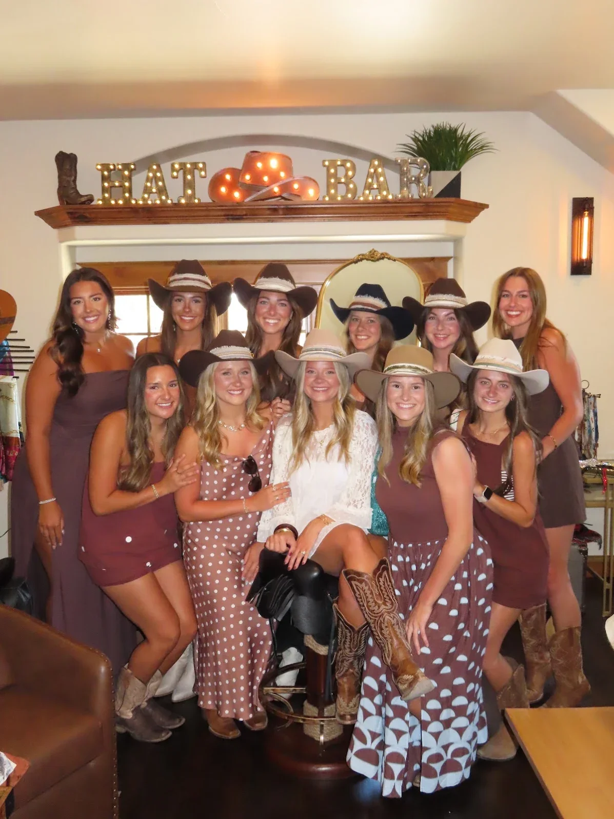 A group of eight women wearing colorful dresses and wide-brimmed hats smiling for a photo indoors, with a green moss wall and neon sign that reads 'Hello there' in the background.