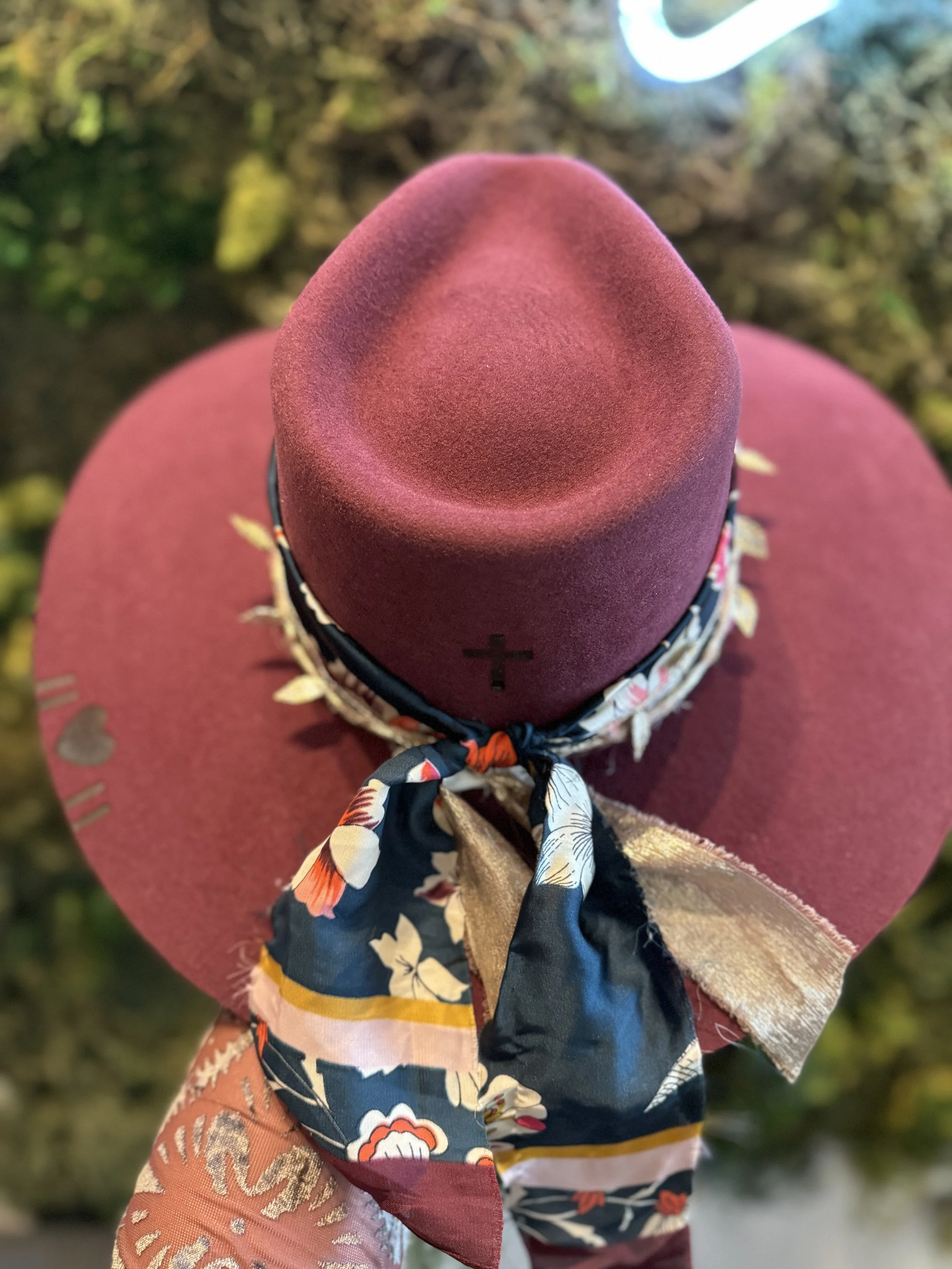 A wide-brimmed maroon hat with a black band and bow, seen from above, with a multicolored scarf tied around the base of the crown, against a blurred outdoors background.