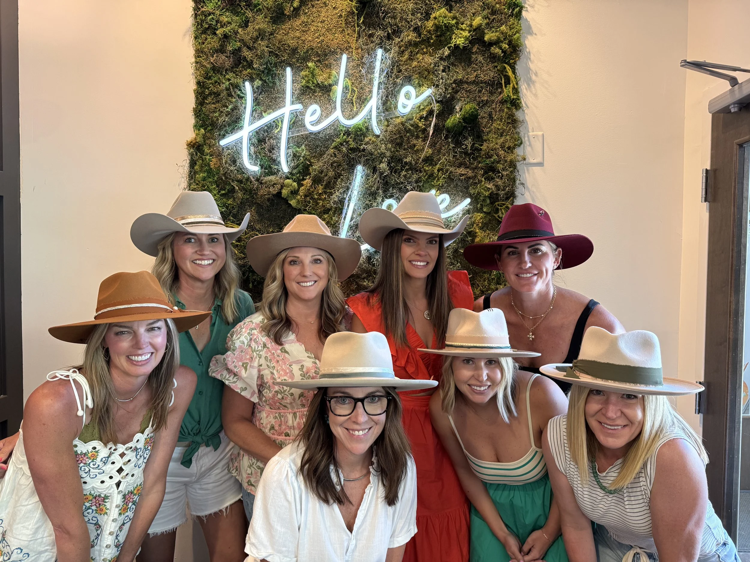 A group of eight women wearing colorful dresses and wide-brimmed hats smiling for a photo indoors, with a green moss wall and neon sign that reads 'Hello there' in the background.