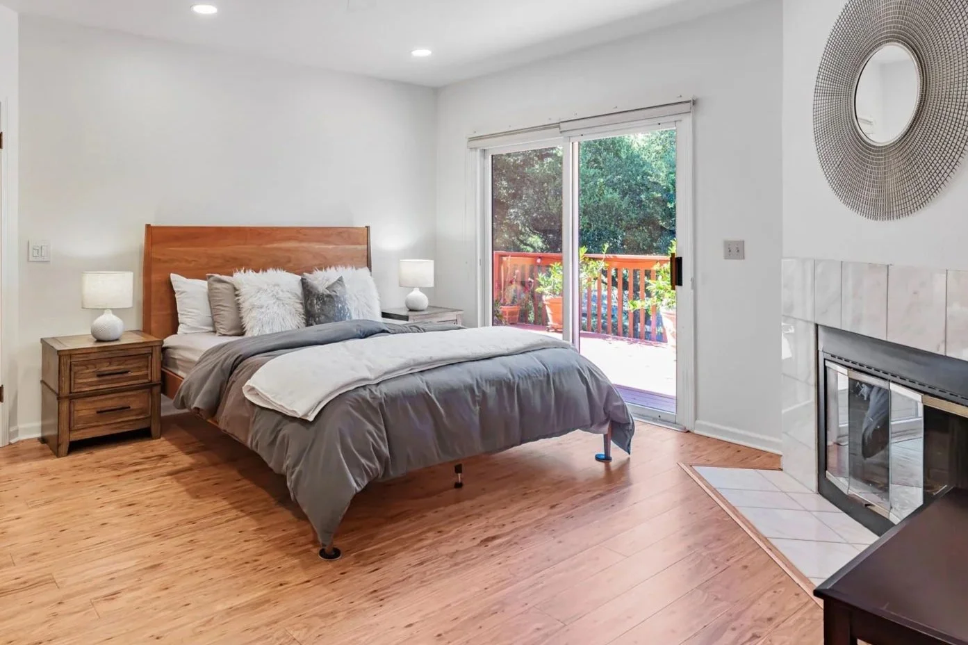 A bedroom with a custom mid-century modern cherry wooden bed, gray bedding, two white lamps on wooden nightstands, decorative pillows, a sliding glass door leading to a patio with potted plants, and a fireplace with a decorative circular mirror.
