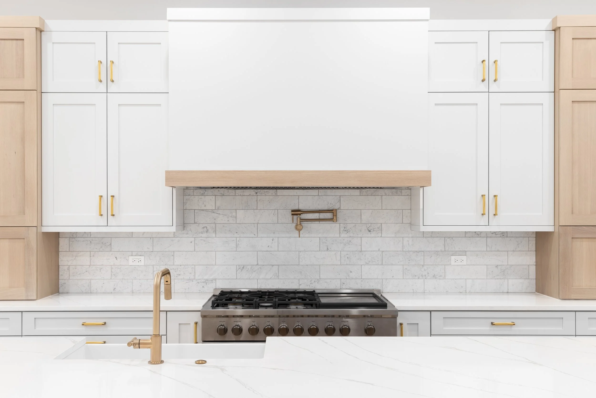 Modern minimalist kitchen with white shaker cabinets, natural wood accents, gold hardware, marble backsplash, and a professional gas range with a pot filler.
