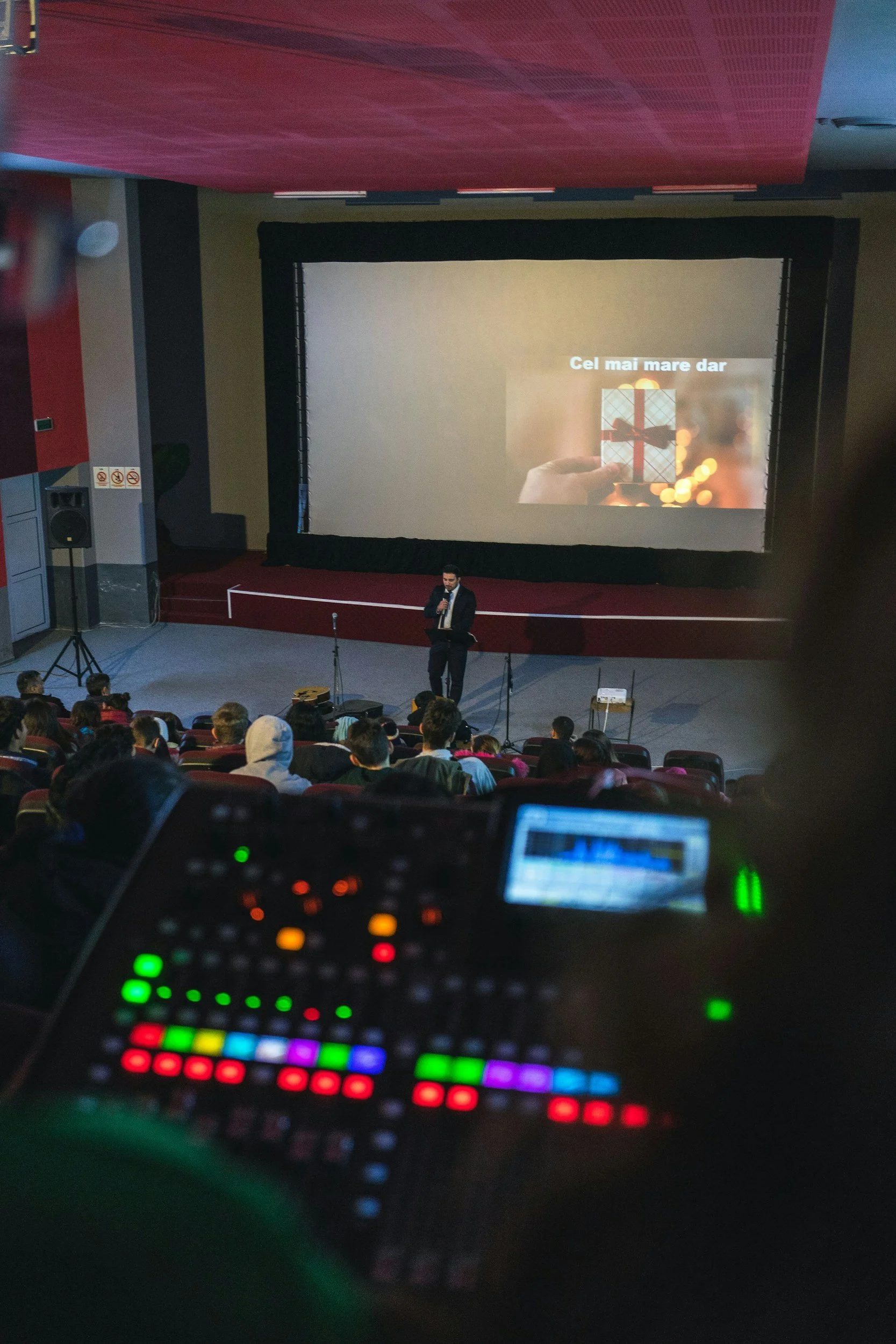 A person speaking on stage in front of an audience in a theater, with a large projection screen showing a gift box and text in Romanian, 'Cel mai mare dar'.