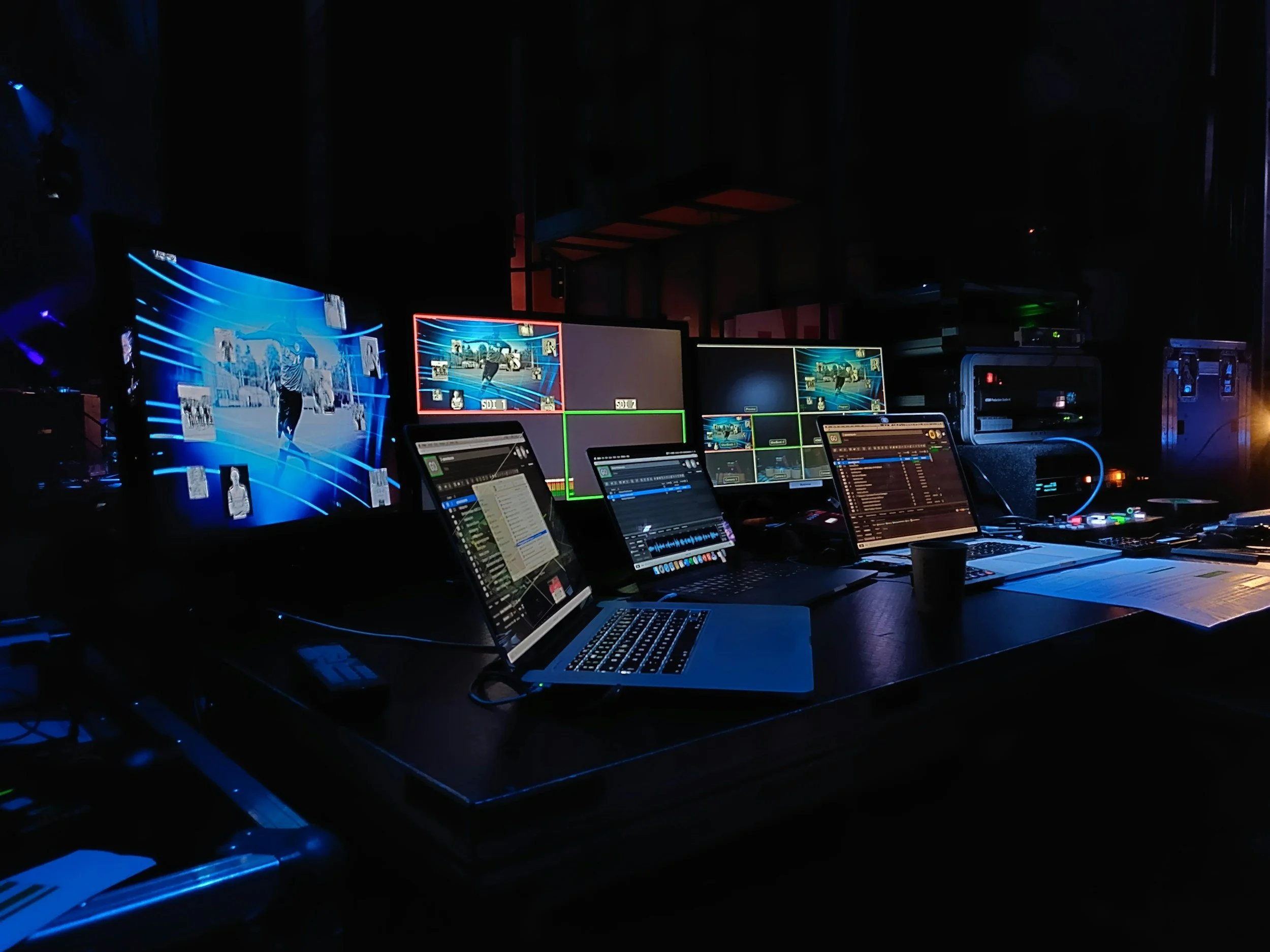 A dimly lit control room with multiple monitors displaying sports and media graphics, laptops, and various electronic equipment.