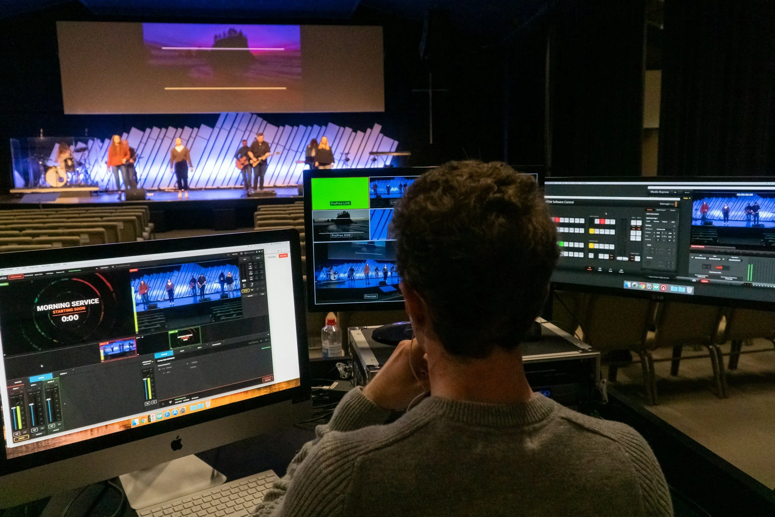 A person working at a control station with multiple monitors, overseeing a live performance of a band on stage in a theater.