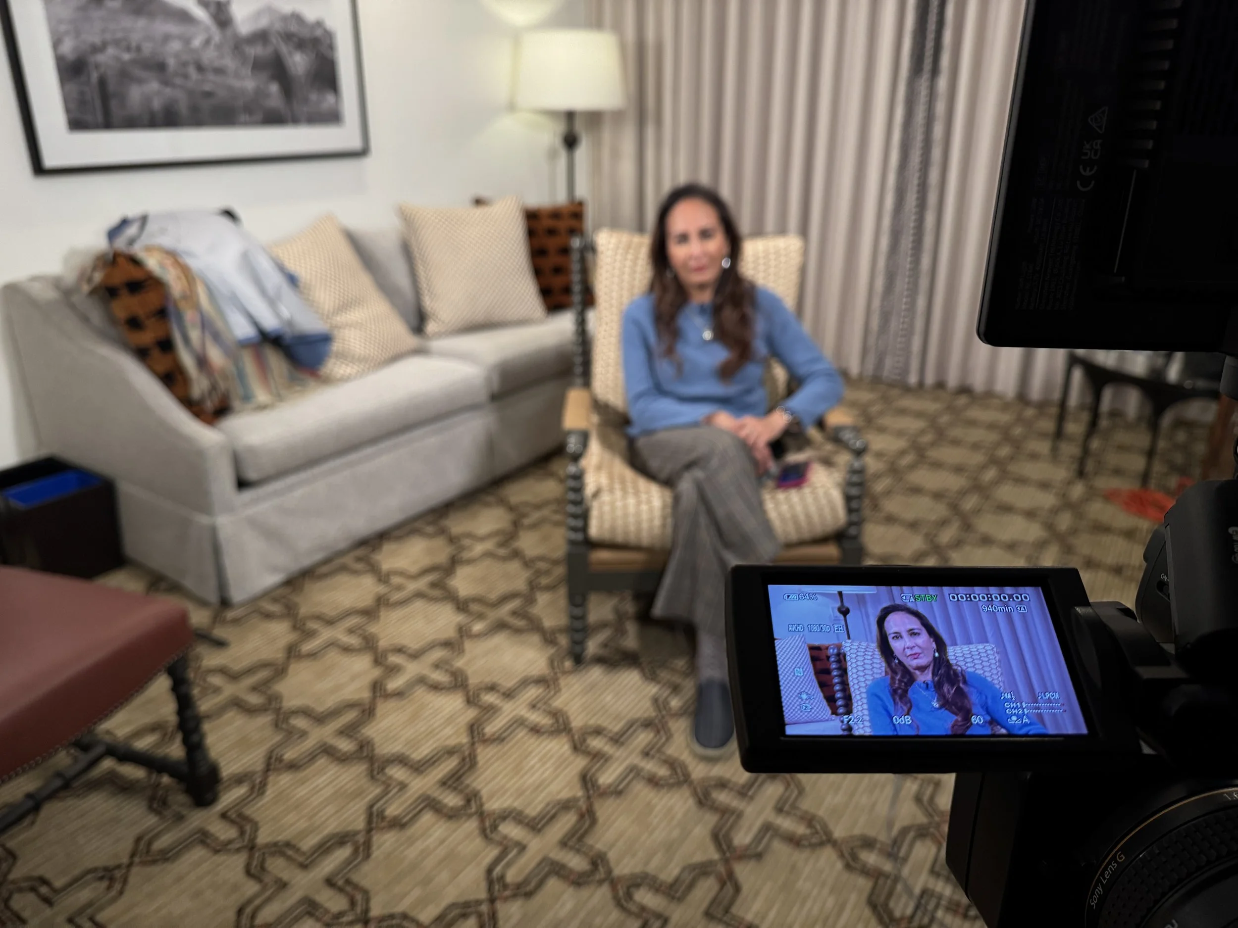 A woman sitting on a patterned armchair in a living room during an interview, with a sofa and curtains in the background, and a video camera filming her.