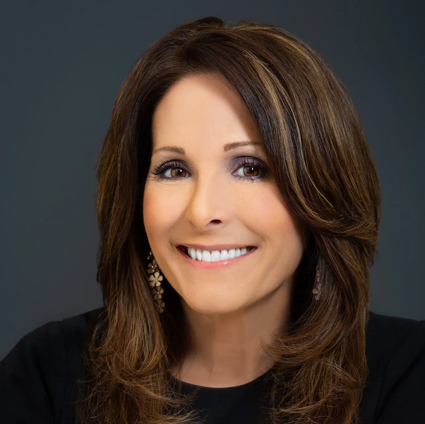 A woman with medium-length brown hair wearing a black top and floral earrings, smiling against a gray background.