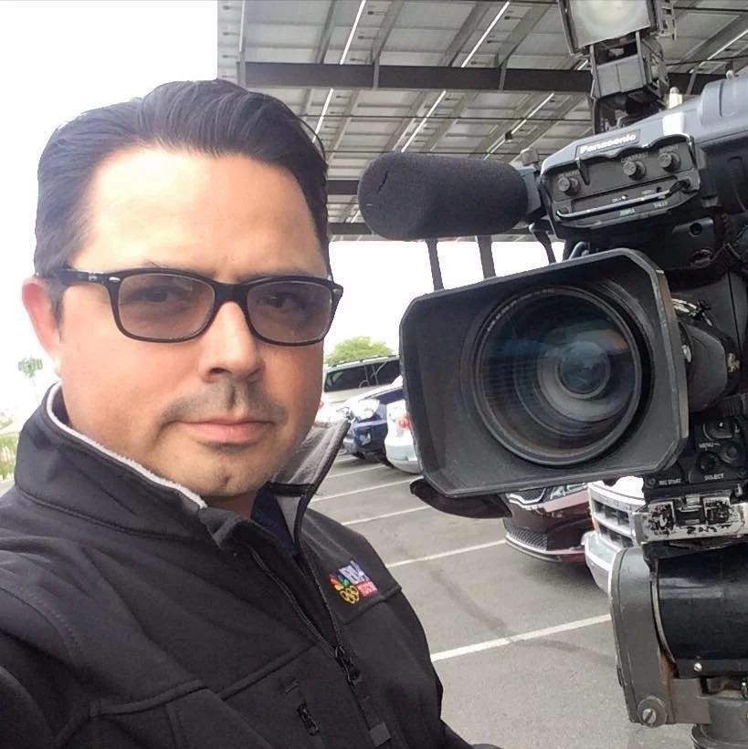 A man wearing glasses and a jacket taking a selfie with a large professional video camera in a parking lot under a metal roof.