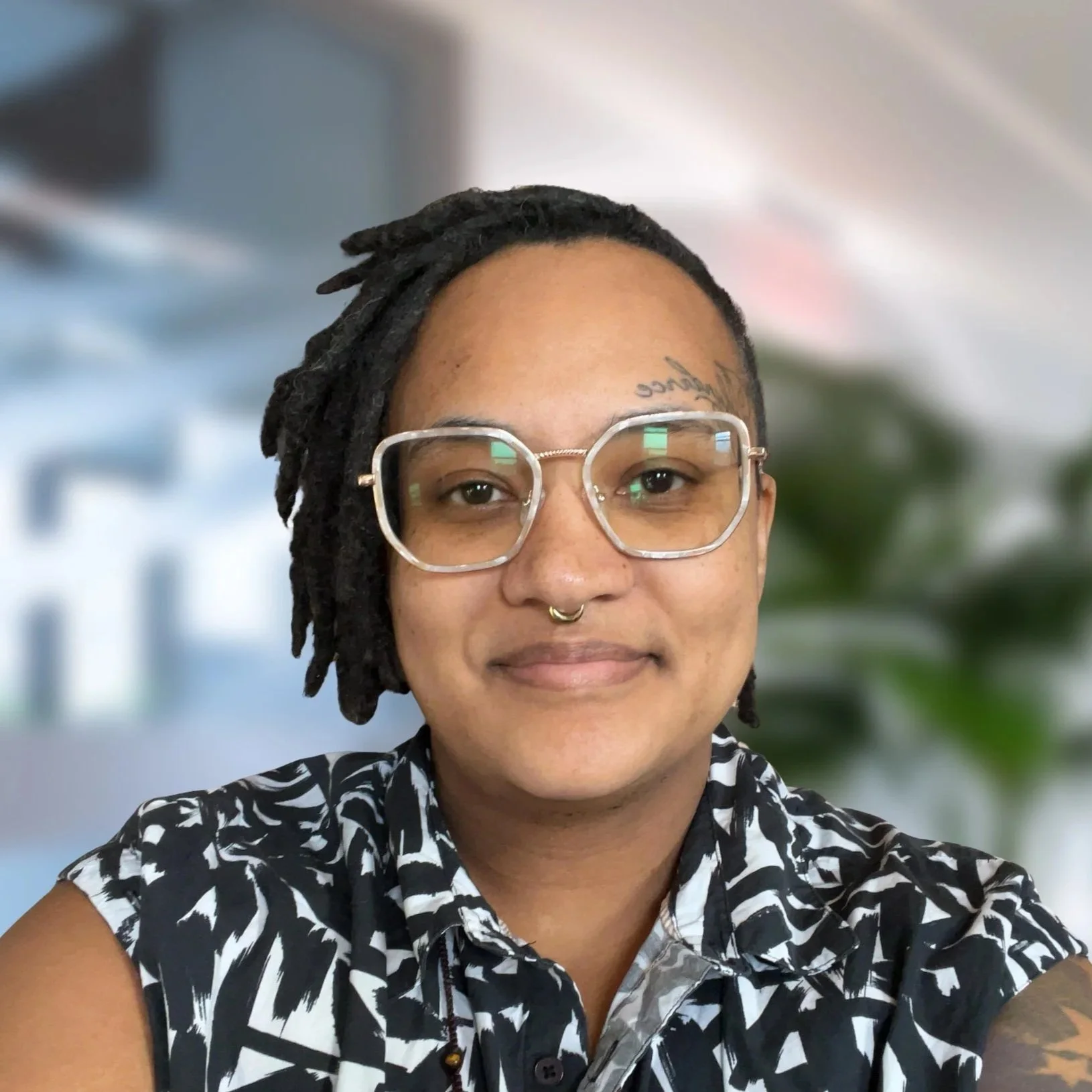 A person with short dreadlocks, wearing glasses, a nose ring, and a black and white patterned shirt, smiling in an indoor setting with blurred background.