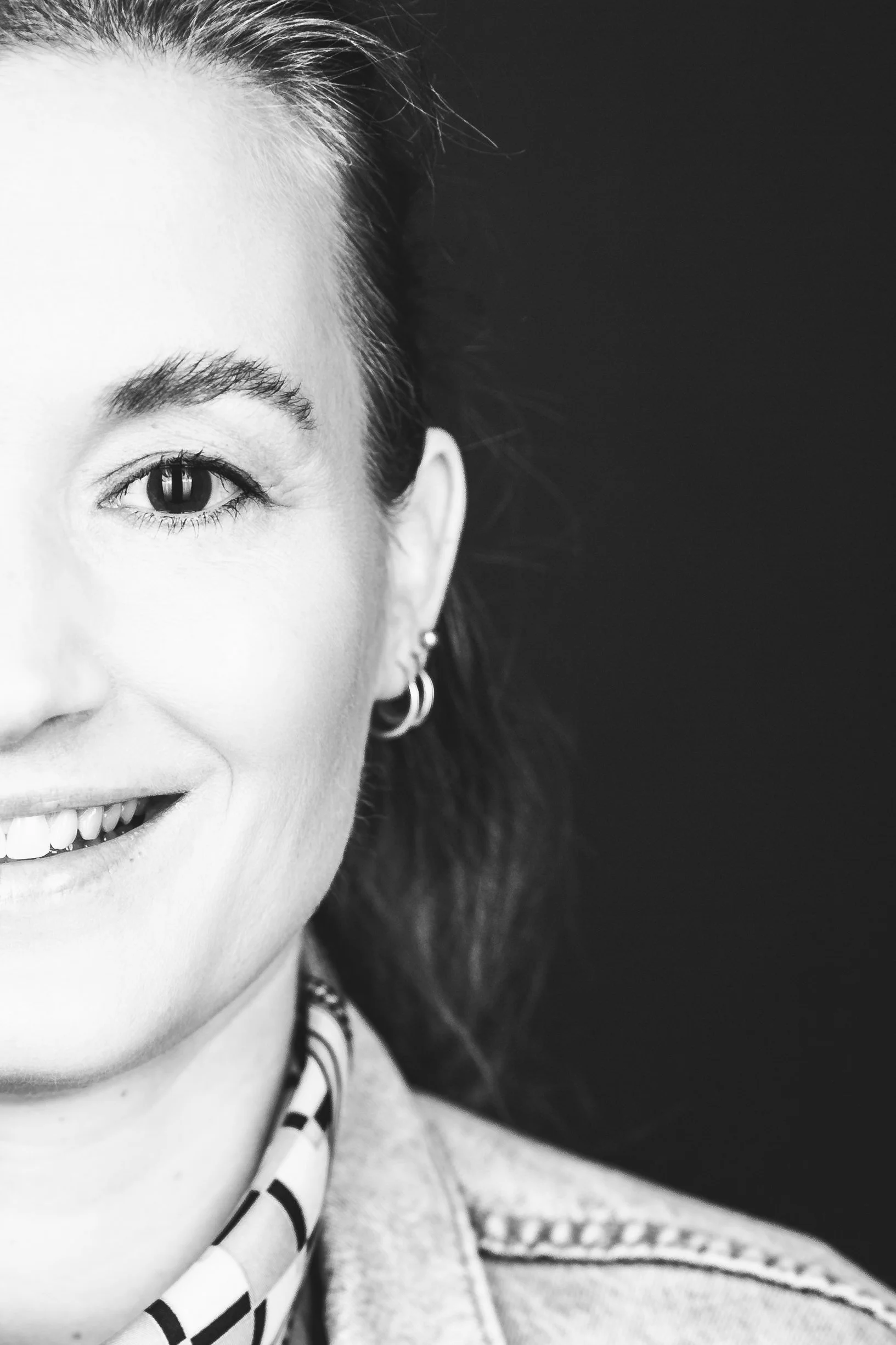 Close-up zwart-wit foto van een lachende vrouw met rechte donker haar, een oorbel en een colbert, voor een donkere achtergrond.