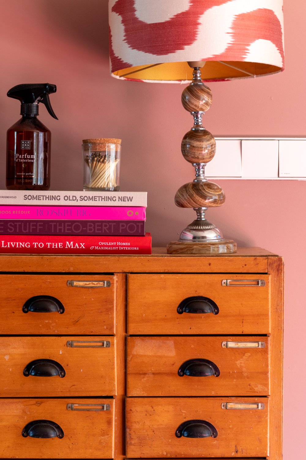 Een houten ladeblok met zes lades, een boekenkast met vier boeken, een rookgels, een glazen pot met lucifers, een tafellamp met een gemarmerde houten voet en een gemarmerde lampenkap, en een roze muur met elektrische schakelaar.