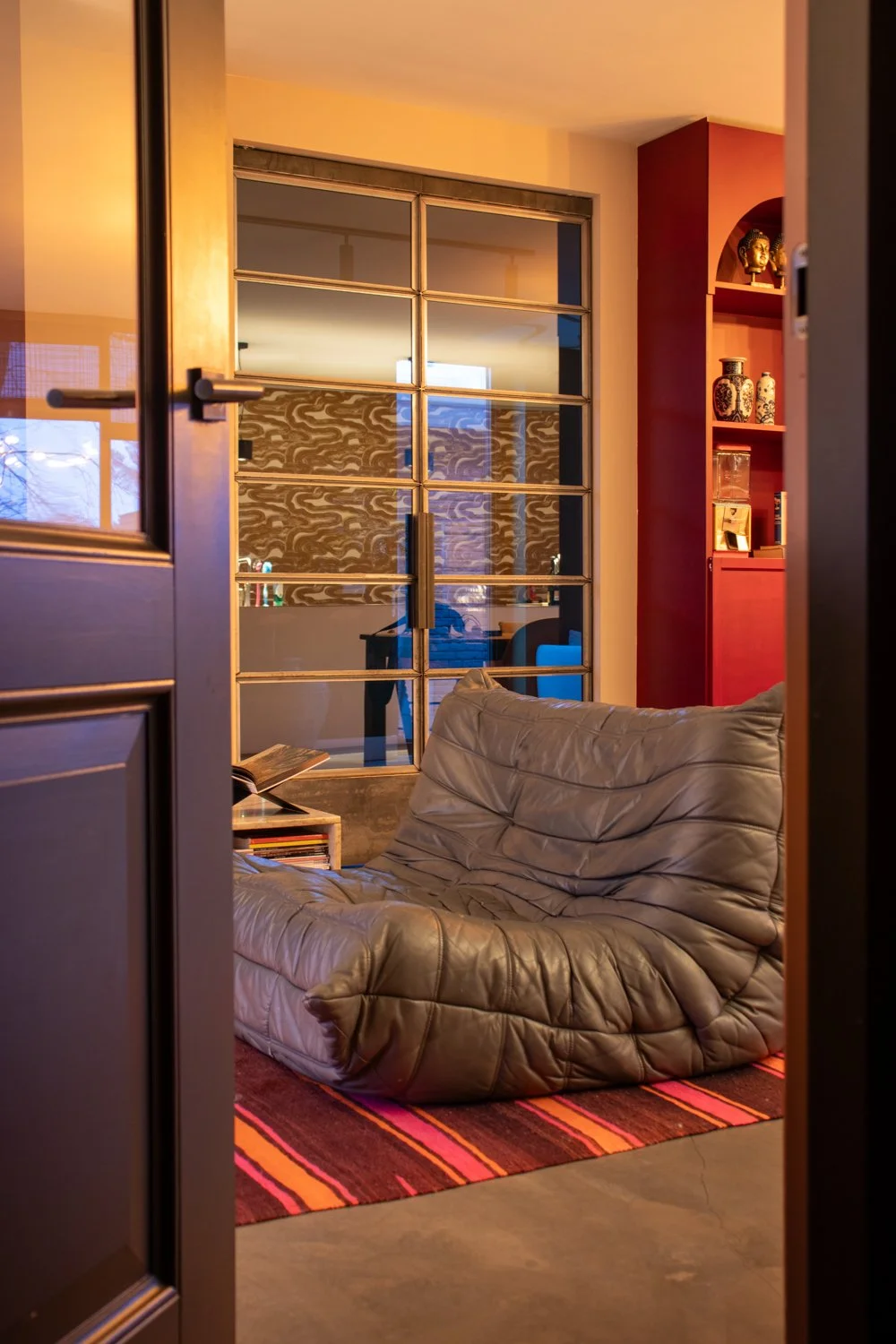 Binnenkant van een woonkamer met een leren fauteuil, rood boekenkast, glazen afscheiding en gedekte ramen.