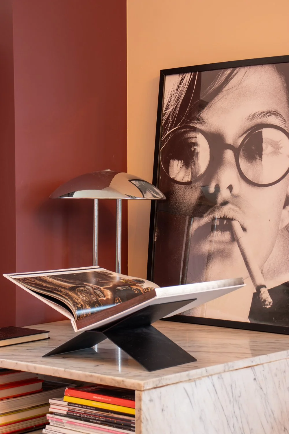 Een boek ligt open op een zwart standaard op een marmeren tafel, er staat een hoekige lamp naast en een grote foto van een vrouw met ronde zwart brillen en een sigaret op de achtergrond.