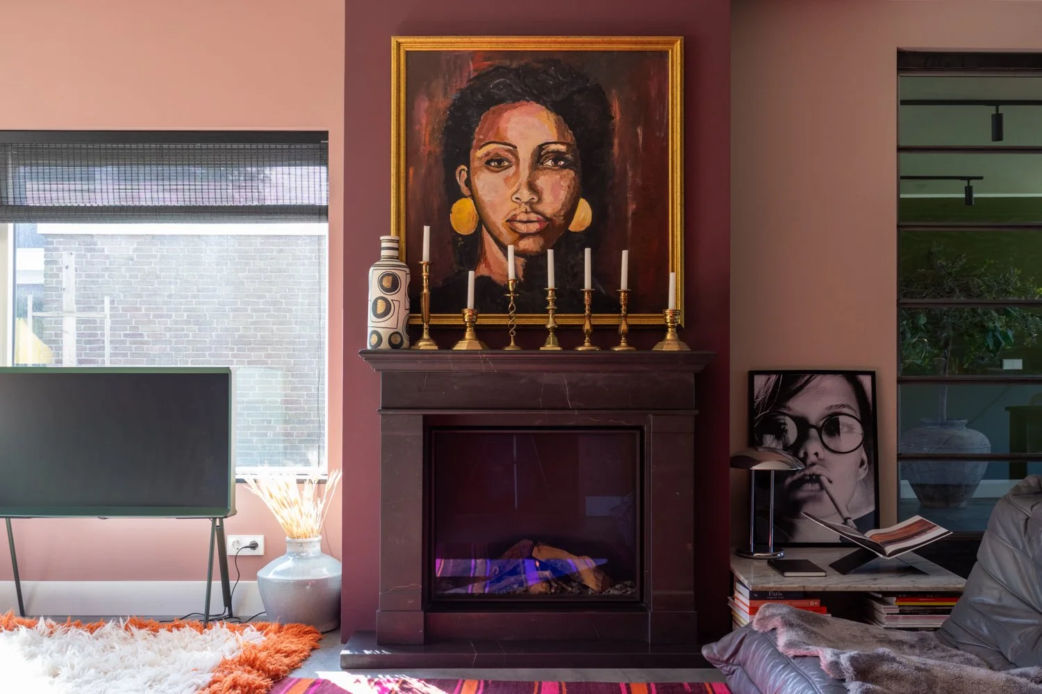 Binnenkijker van een woonkamer met een zwarte open haard, een groot portret van een vrouw met grote oorbellen op de muur, een tv, een vlammentafel met een boek, een poster en een lamp, een vloerkleed en een venster met jaloezieën.