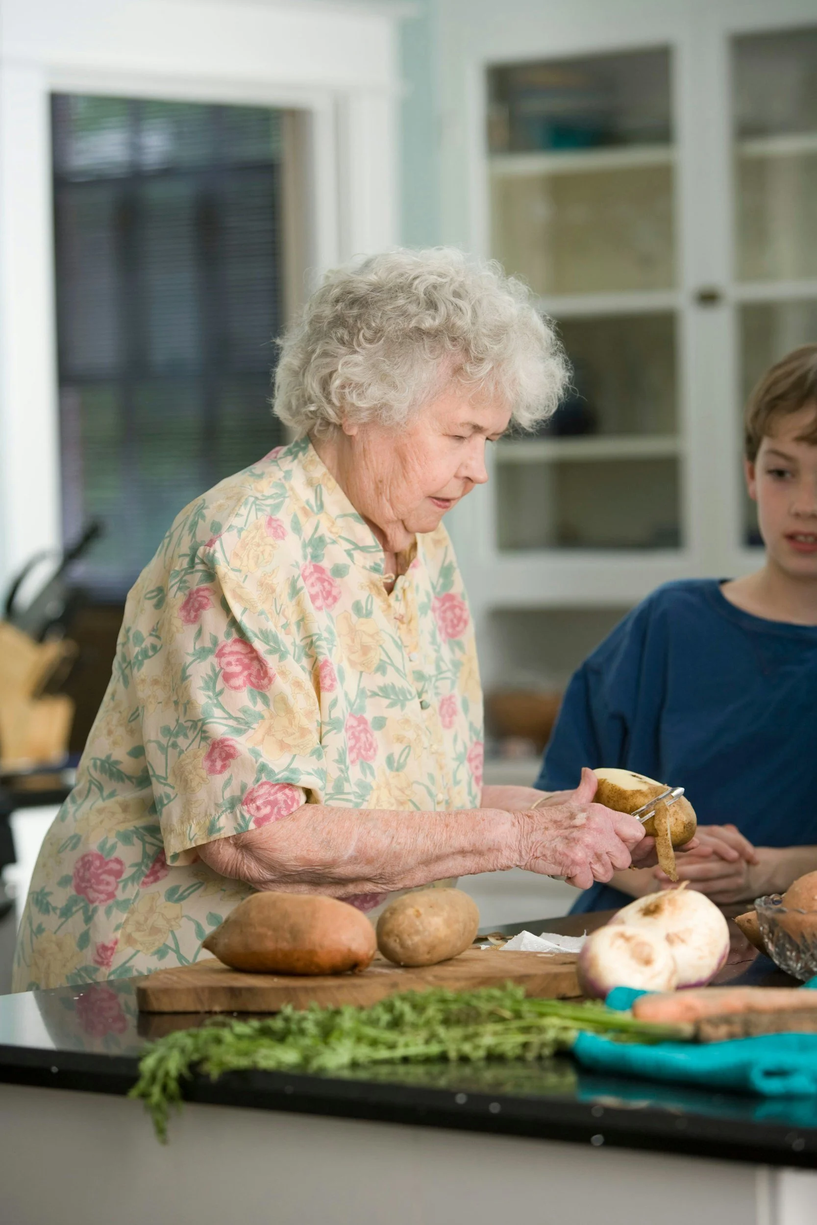 An elderly woman with gray curly hair slicing a potato in a kitchen, with a young boy beside her and various vegetables on the counter.
