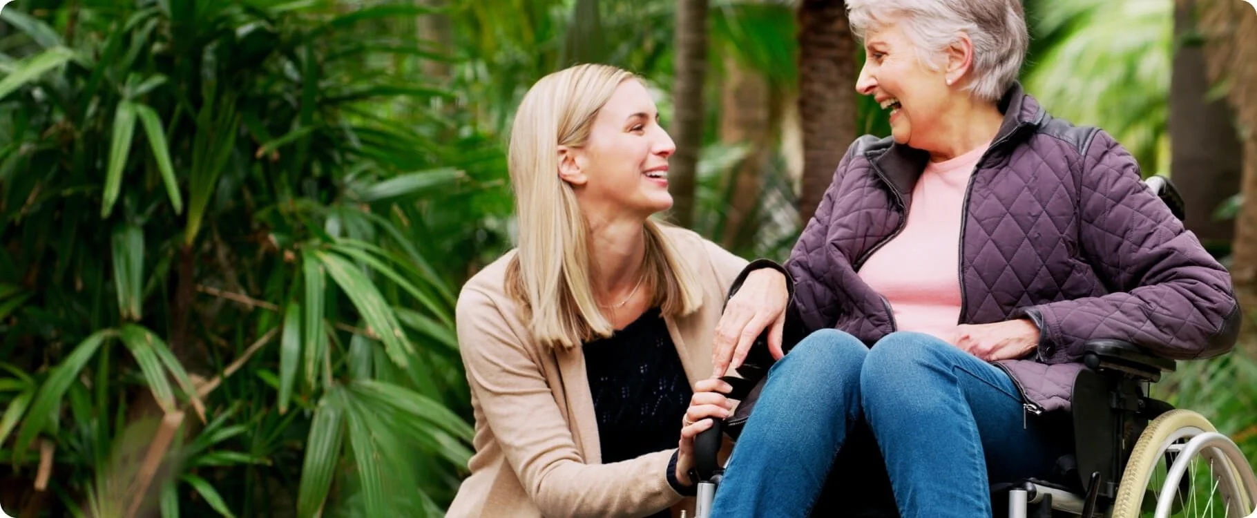 A young woman helps an elderly woman in a wheelchair smile and laugh outdoors in a lush green garden.