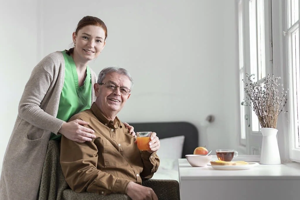 A young woman in medical scrubs standing next to an elderly man in a brown shirt, sitting in a chair, holding a glass of orange juice in a bright room with windows, a table with a bowl of fruit, a cup of tea, and a vase with dried flowers.