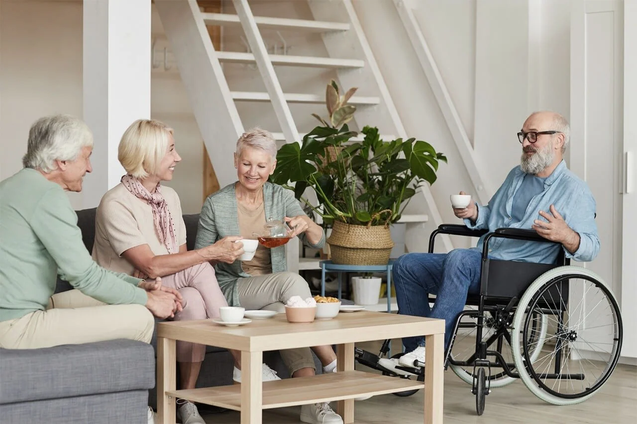 Four elderly adults, one in a wheelchair, enjoying tea and snacks together in a cozy living room.