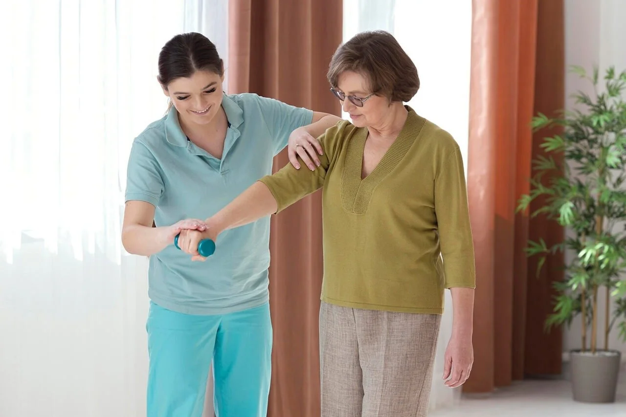 Nursing assistant helping an elderly woman exercise with a light resistance band in a bright room.
