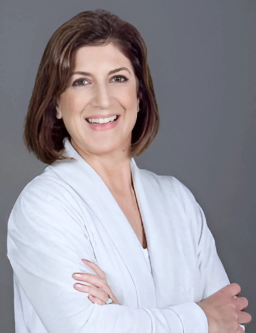 A smiling woman with shoulder-length brown hair, wearing a white top, standing against a gray background.