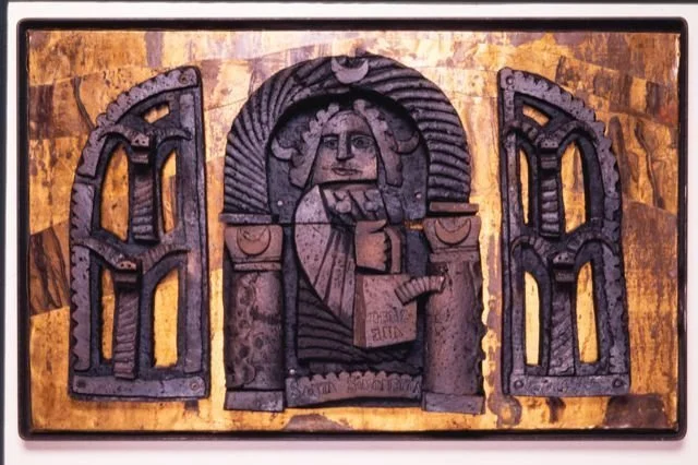 Clay sculpture with a human figure, surrounded by arches and decorative elements, on a textured background.