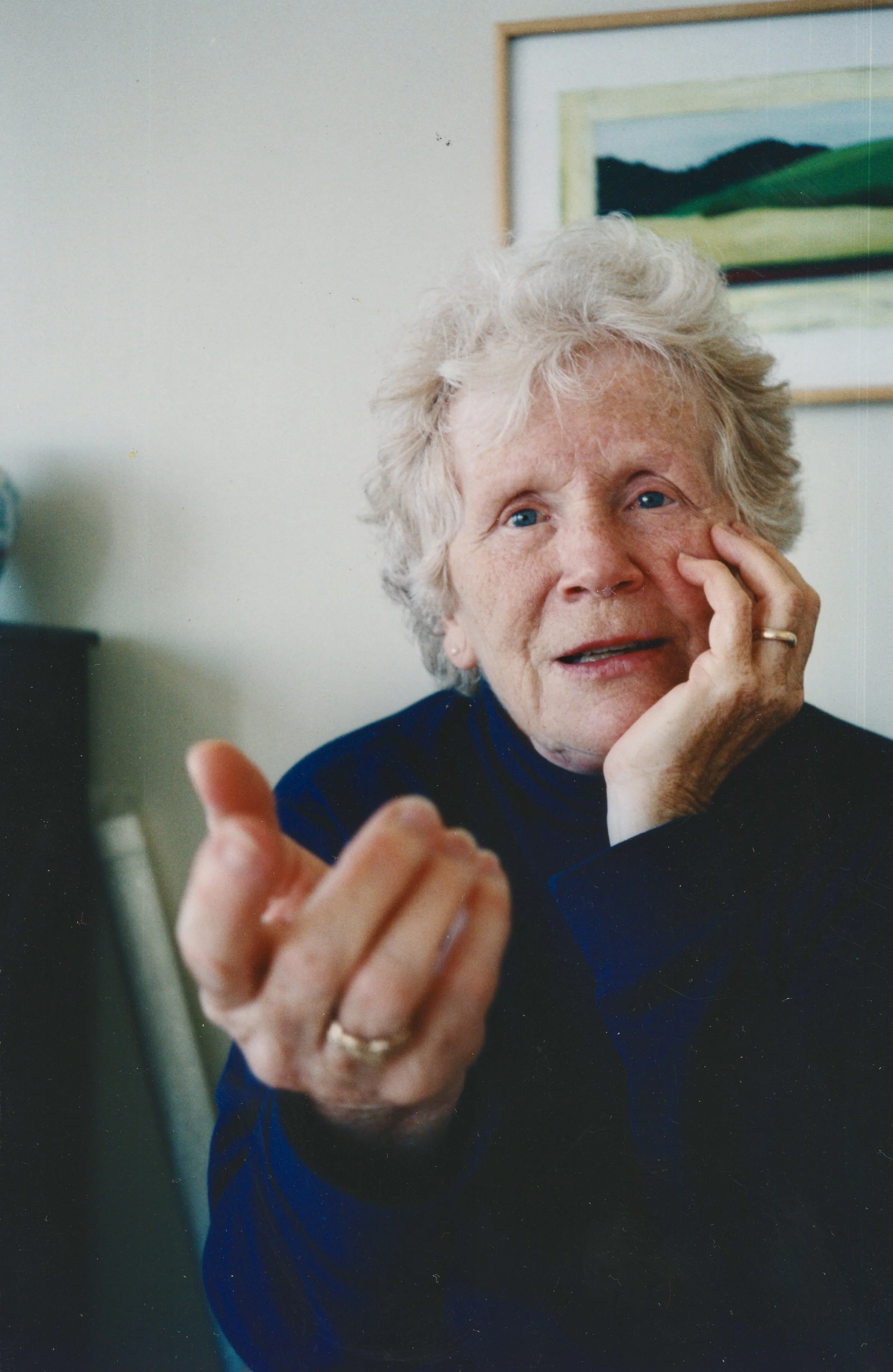 An elderly woman with curly gray hair and blue eyes, wearing a navy blue top, appears to be engaged in a conversation or explaining something. She rests her face on her left hand while her right hand is extended forward. There is a painting of green hills hanging on the wall behind her