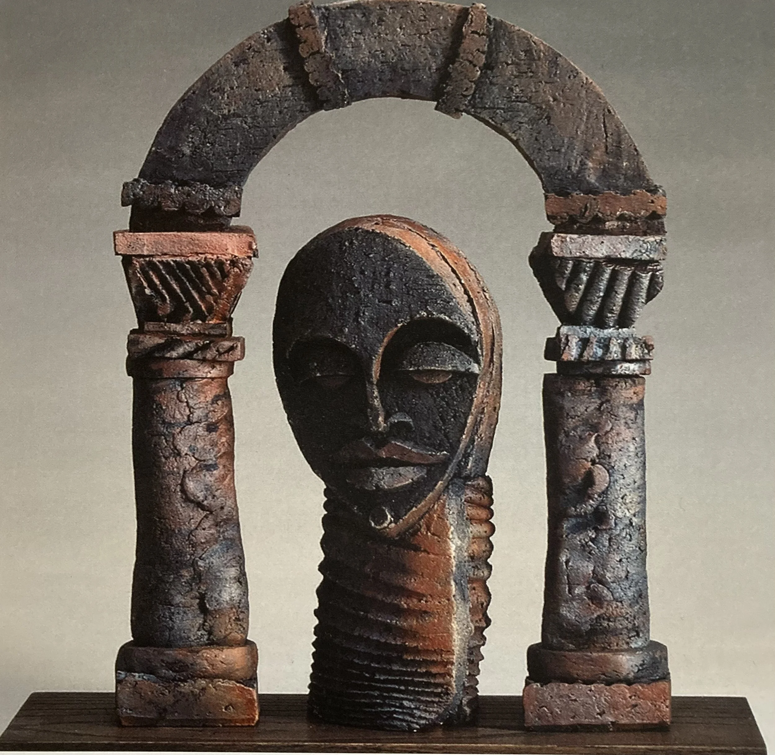 Clay sculpture featuring a human face framed by two columns and an arch, made of rust-colored material with carved details.