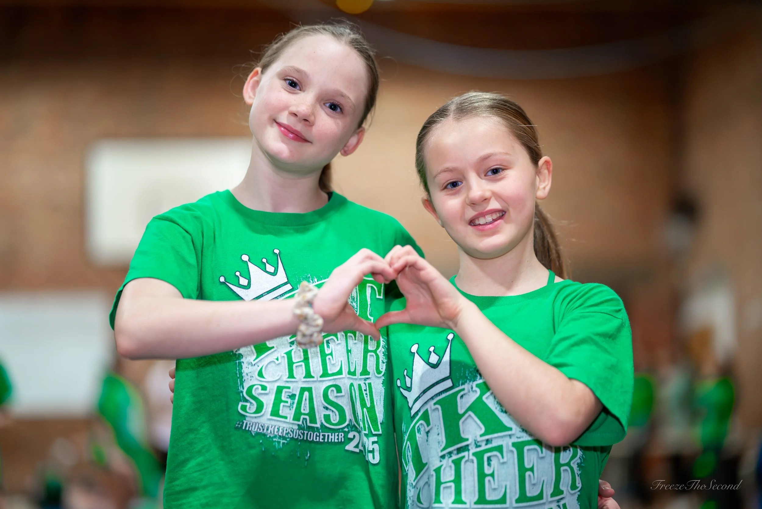 Two young girls with brown hair smiling and forming a heart shape with their hands, wearing matching green cheer season shirts.