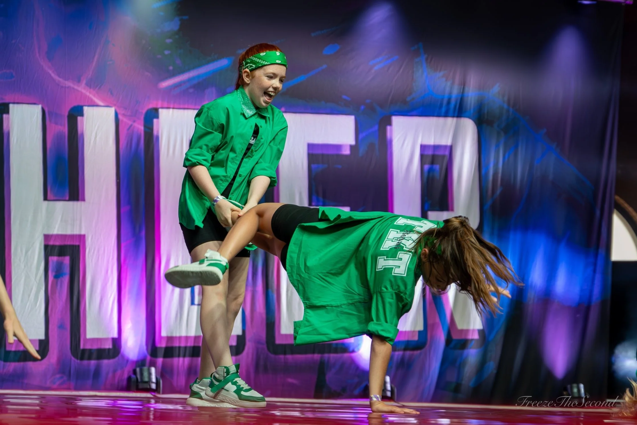 Two girls in green jackets, one standing and one crawling, performing on a stage with a colorful backdrop.