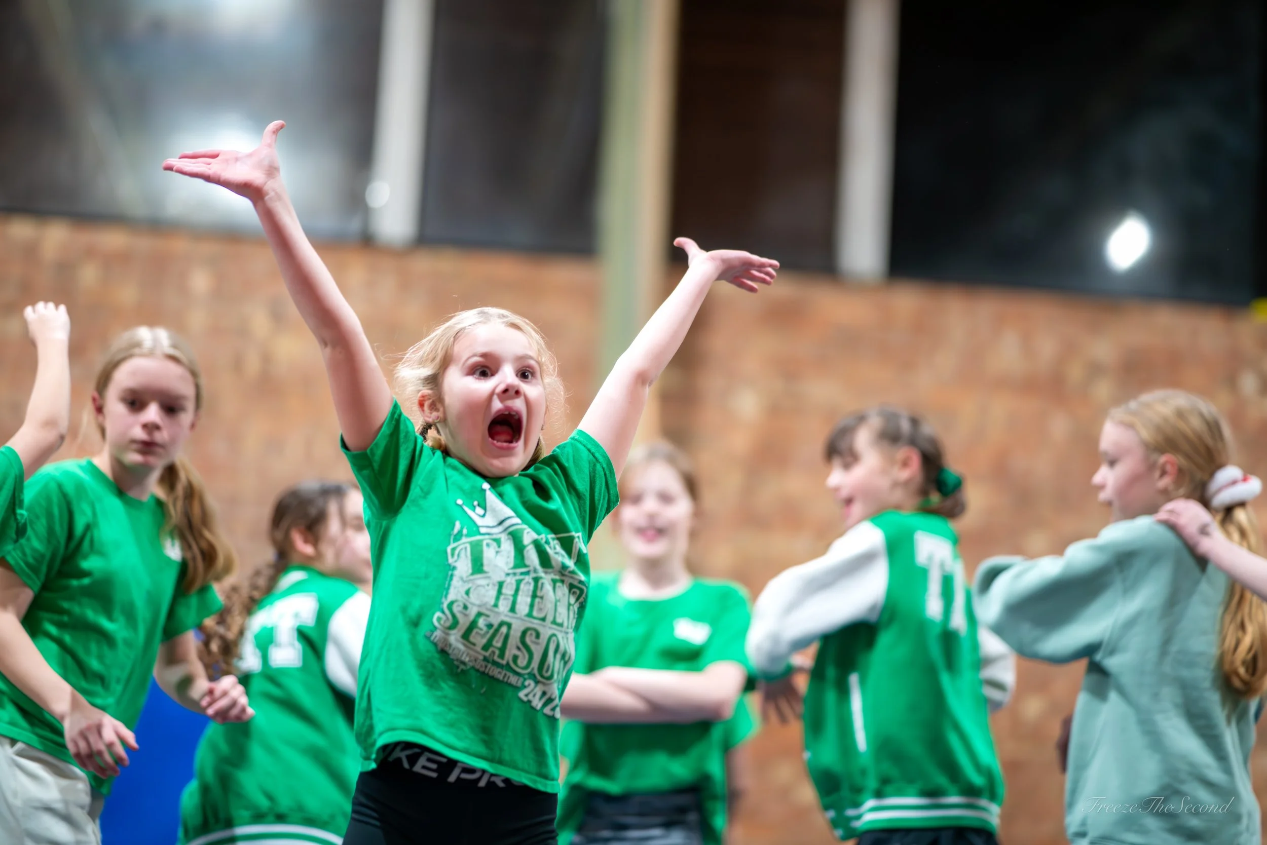 Group of young girls in green sportswear participating in a team activity indoors, with one girl expressing excitement or surprise, and others listening or waiting.