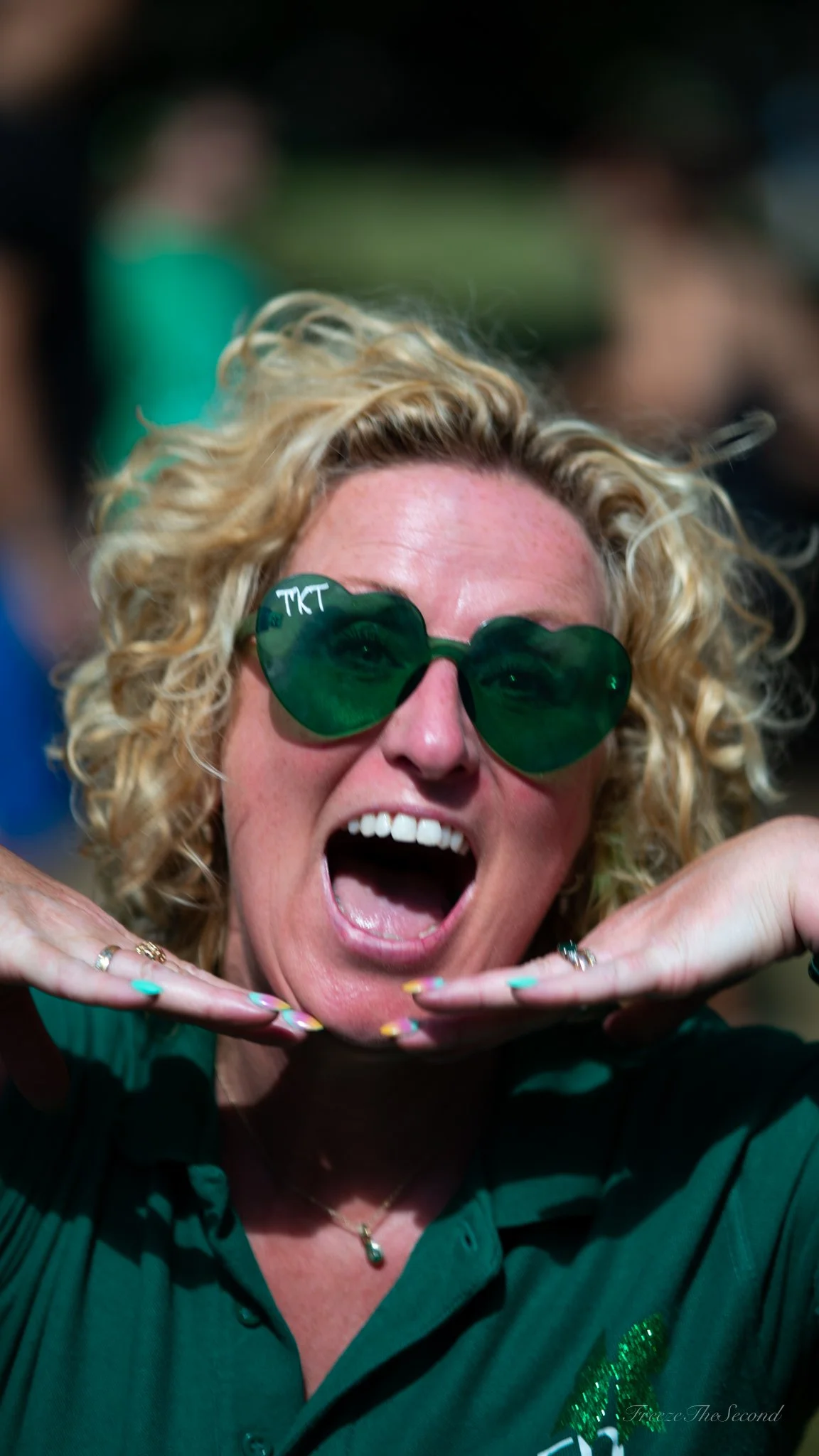 A woman with curly blonde hair, wearing green heart-shaped sunglasses, making a playful gesture with her hands under her chin and smiling with her mouth open. She is wearing a green shirt and appears to be at an outdoor event.