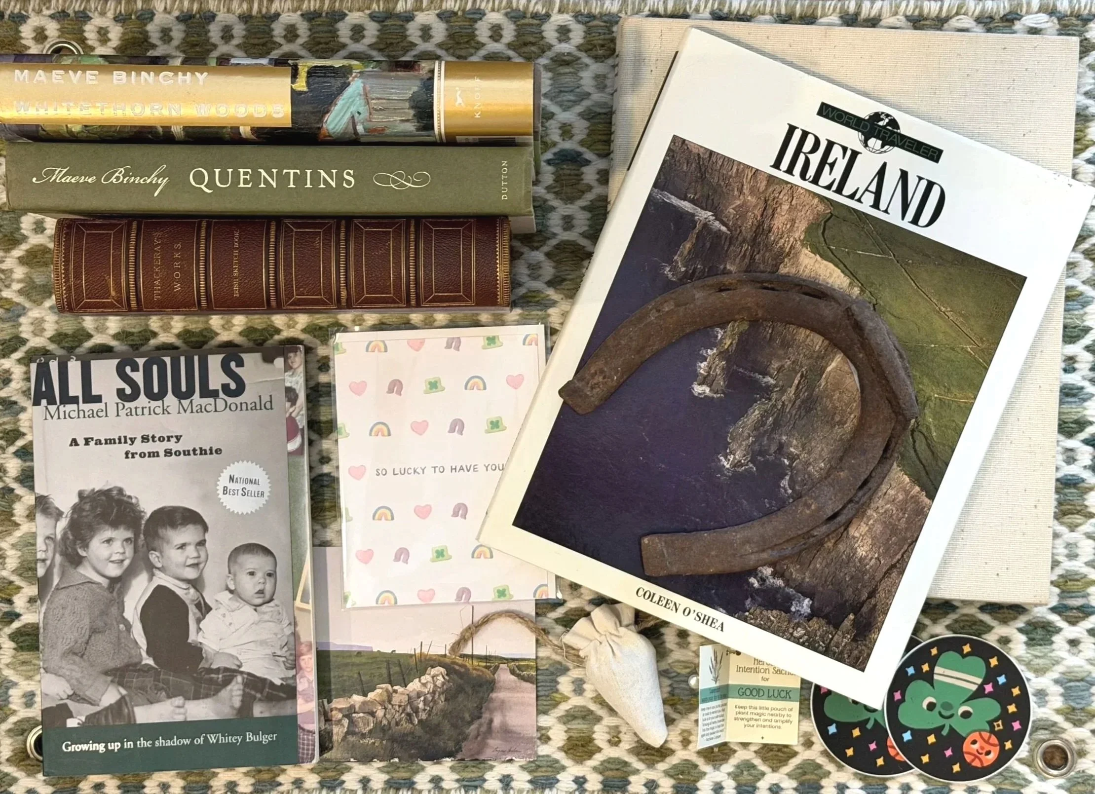 A collection of books about Ireland and family stories, a horseshoe, a small framed picture of a countryside road, a shell, stickers, and a gift card, laid out on a woven mat.
