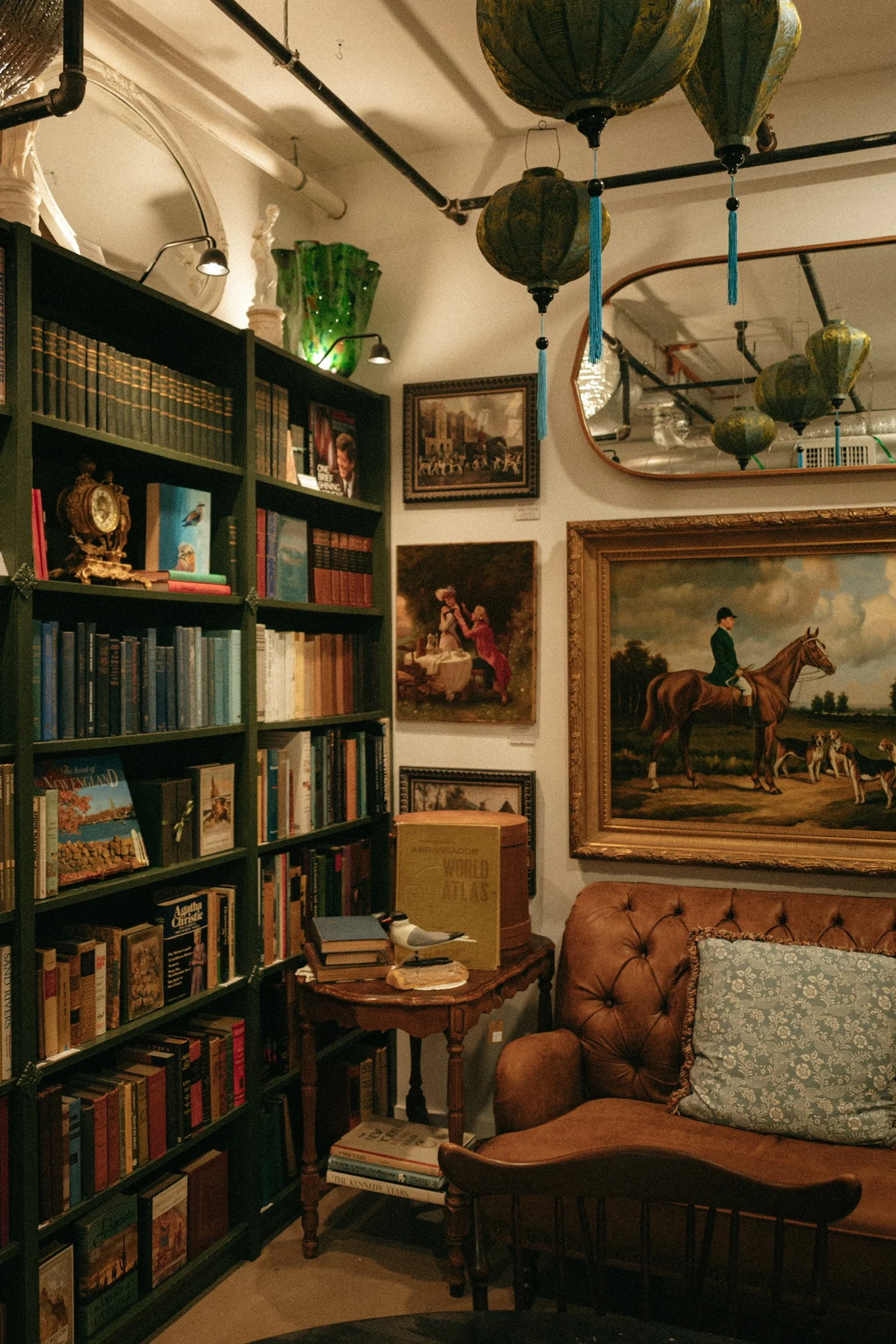 A cozy room with a green bookshelf filled with books, framed paintings on the wall, hanging green lanterns from the ceiling, a brown leather tufted sofa with a patterned throw pillow, a small round wooden side table with books, a bird sculpture, and a large framed painting of a person riding a horse.